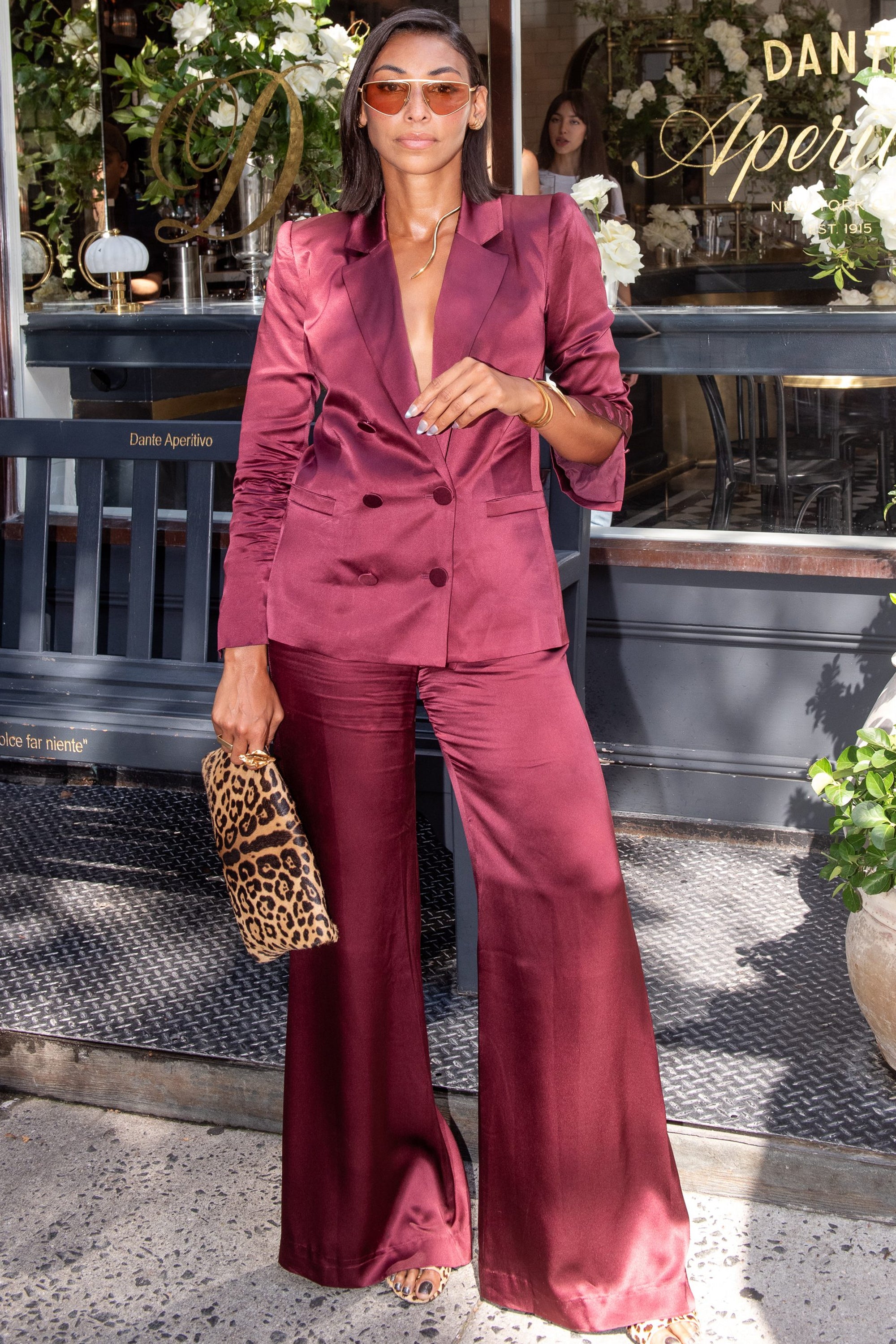 A woman in a shiny burgundy suit and sunglasses stands outside a café, holding a leopard print clutch and wearing heeled sandals. White flowers frame the dark wood café entrance in the background, adding elegance to the scene.