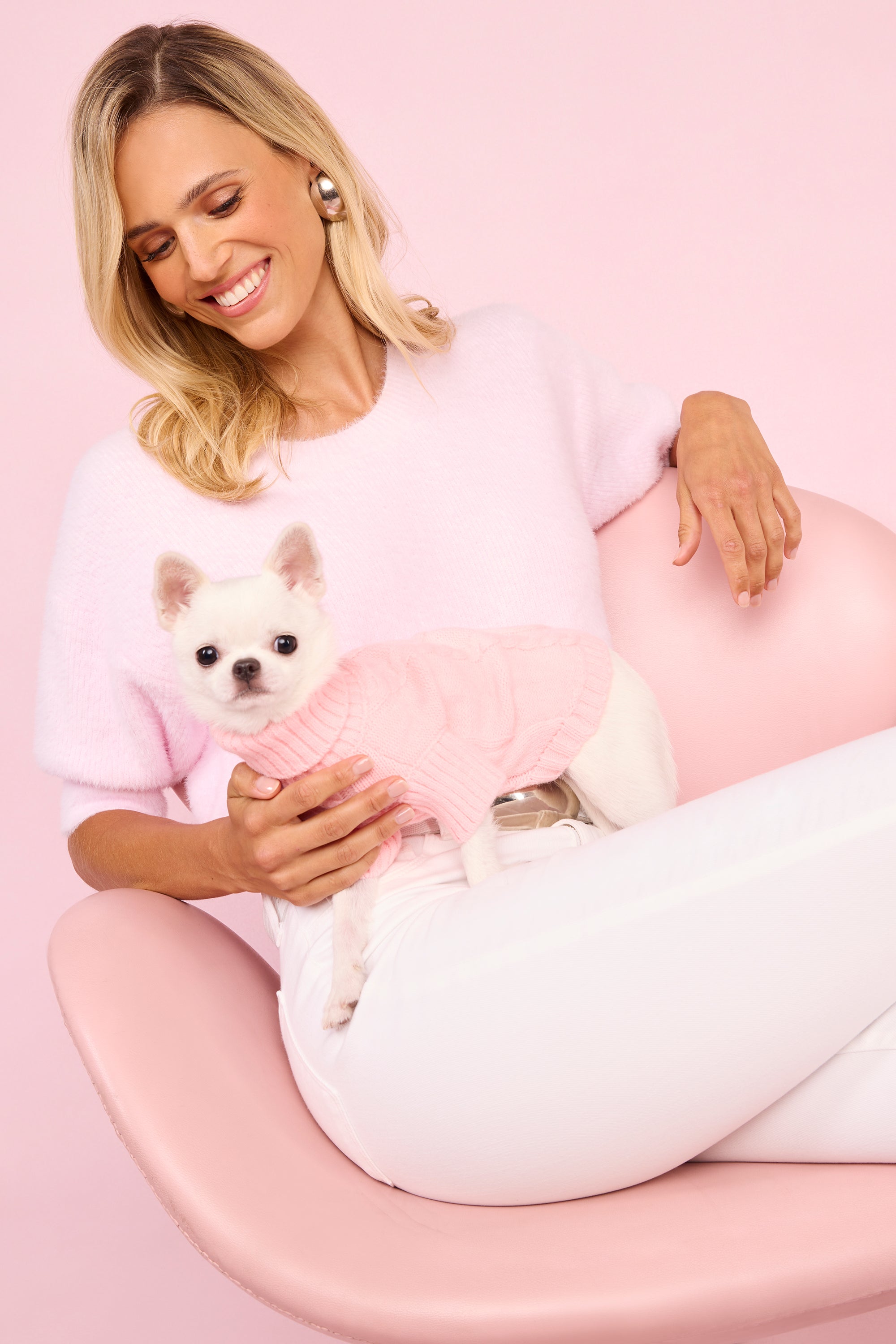 A smiling woman in a pink sweater and white pants sits on a pink chair, holding a small white dog dressed in a cozy pink sweater, set against a vibrant pink background.