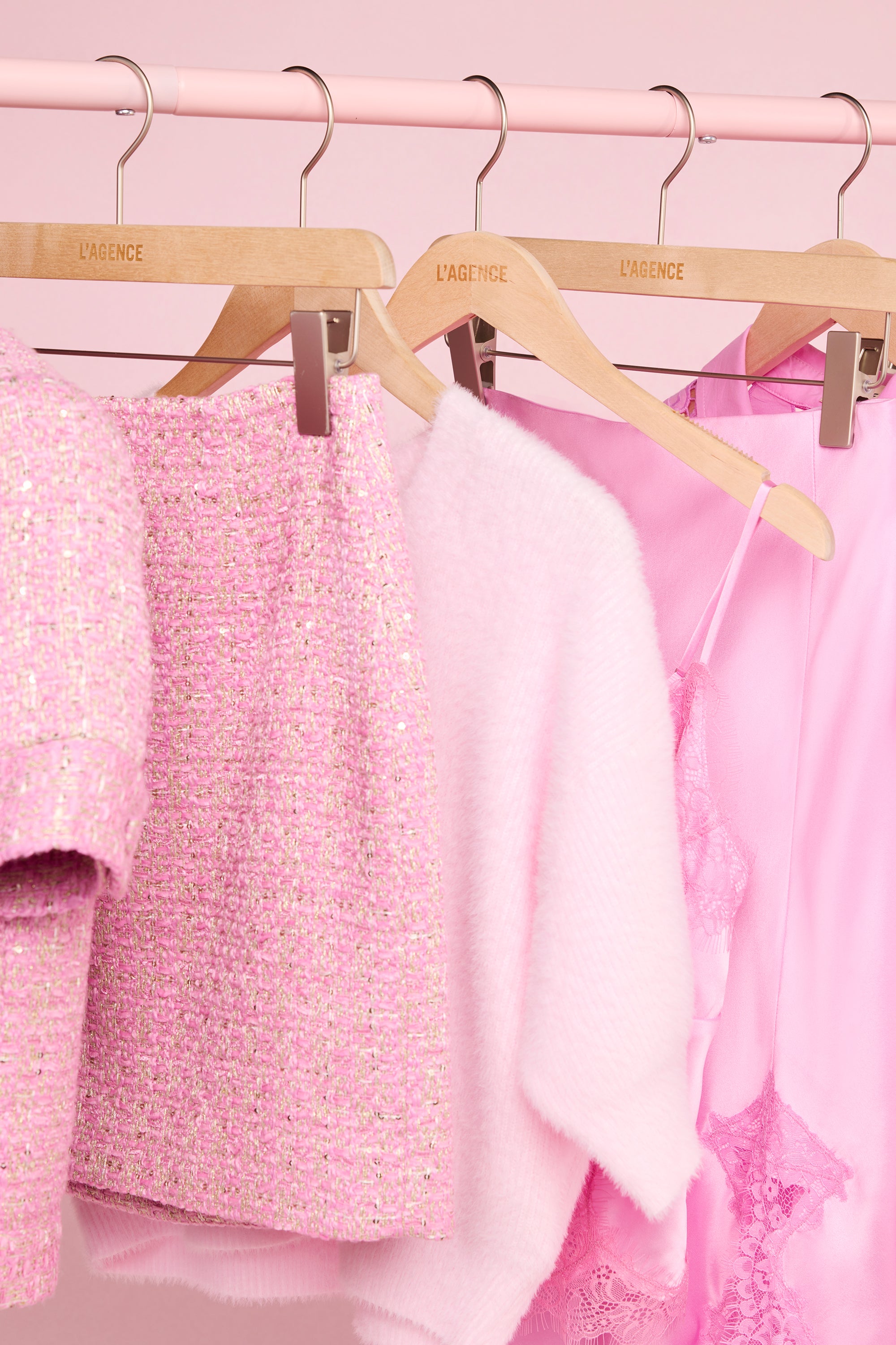 Four pink garments, including a tweed skirt, fuzzy top, and lace-trimmed dress, hang on wooden hangers from a pastel pink clothing rack against a pink background.