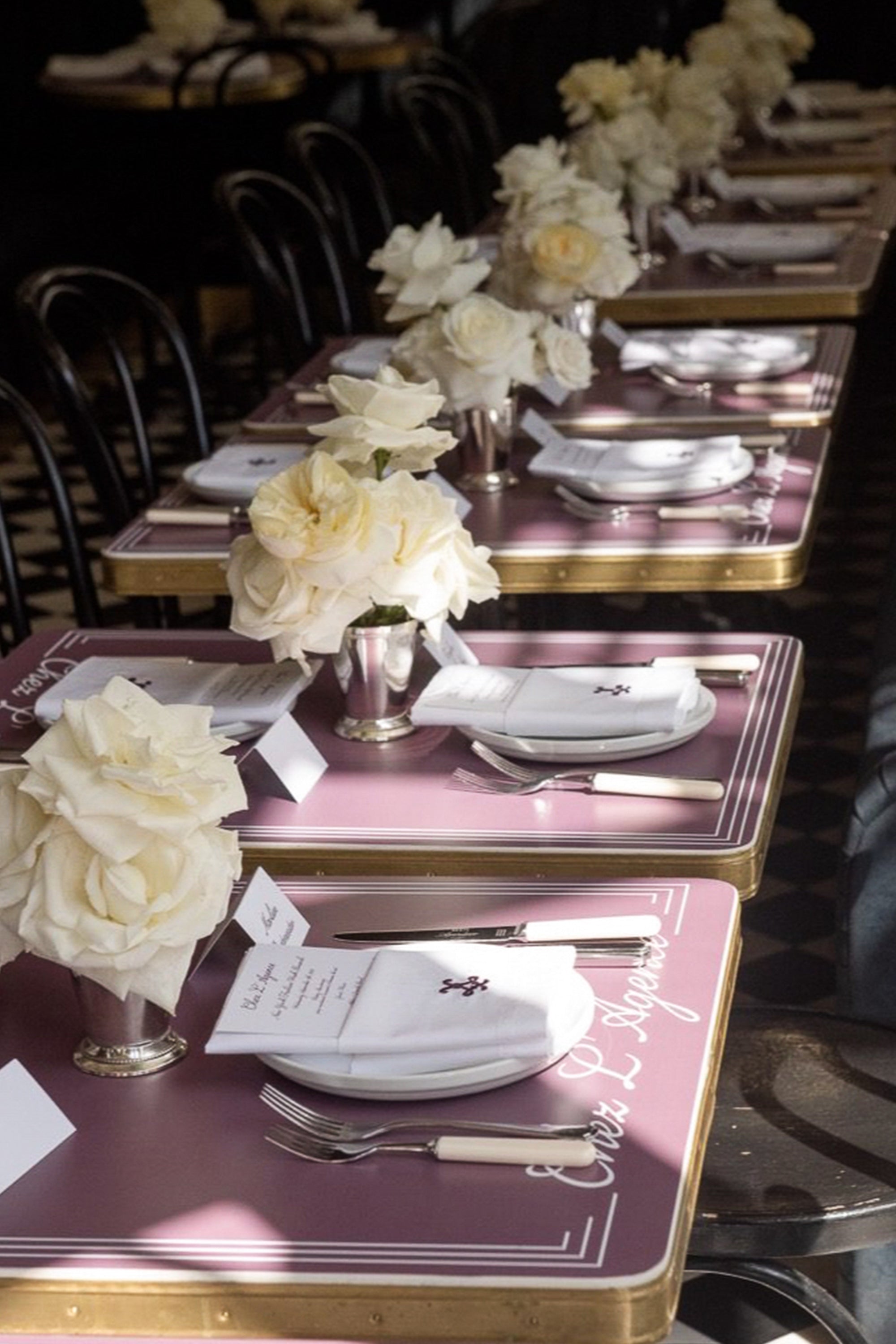 Rows of tables with purple placemats, white napkins, menus, silverware, and small vases holding white roses are arranged neatly in a softly lit restaurant atmosphere.