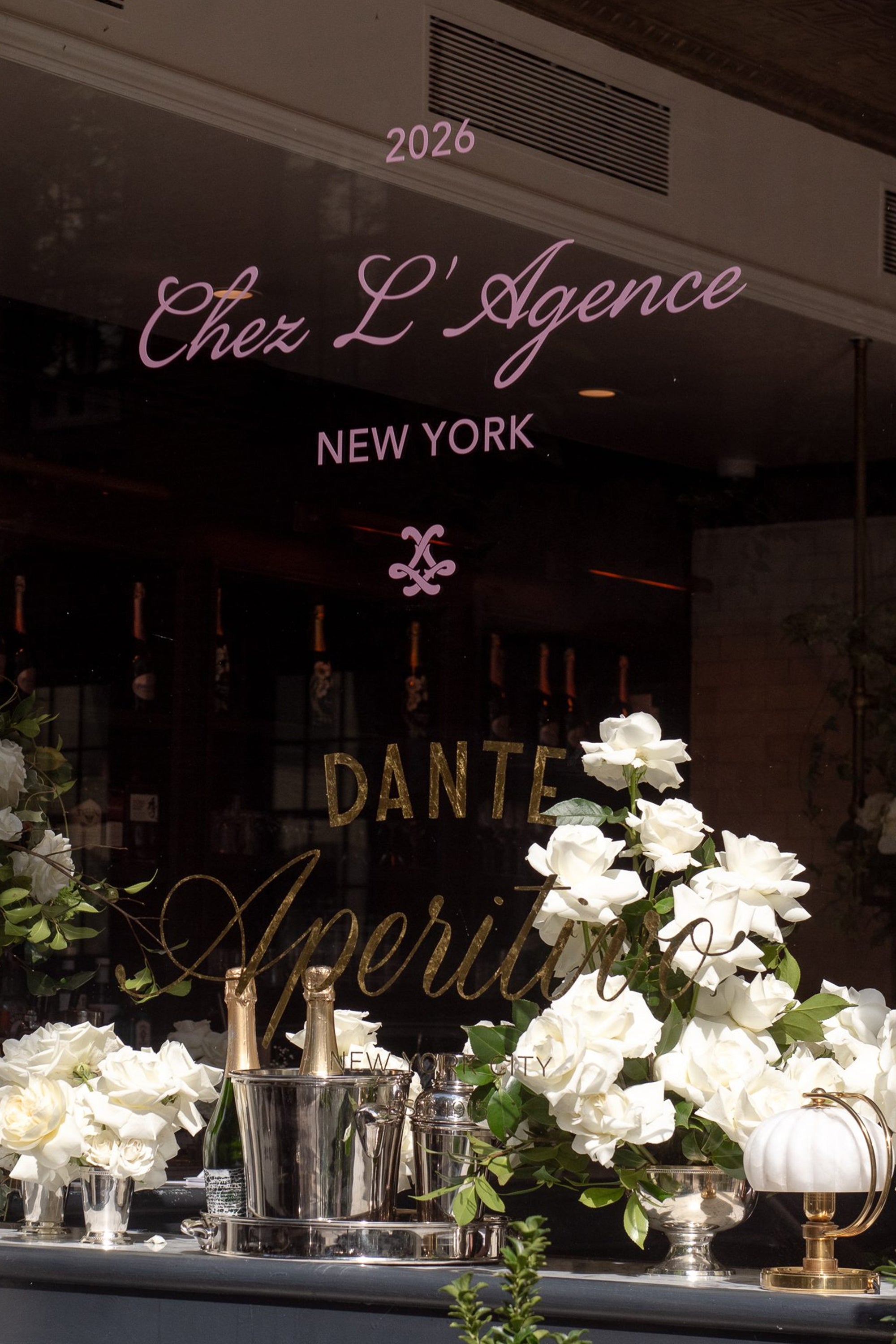 A restaurant window displays floral arrangements, bottles of champagne, and signage reading Chez LAgence New York and Dante Aperitivo, with reflections and elegant aperitivo-inspired decor.