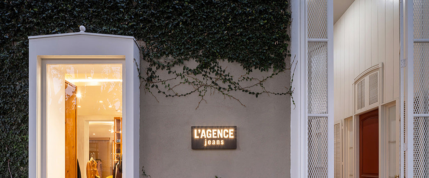 Exterior of a building with ivy-covered walls, a lit LAGENCE jeans sign, and a large open doorway displaying denim clothing racks inside.