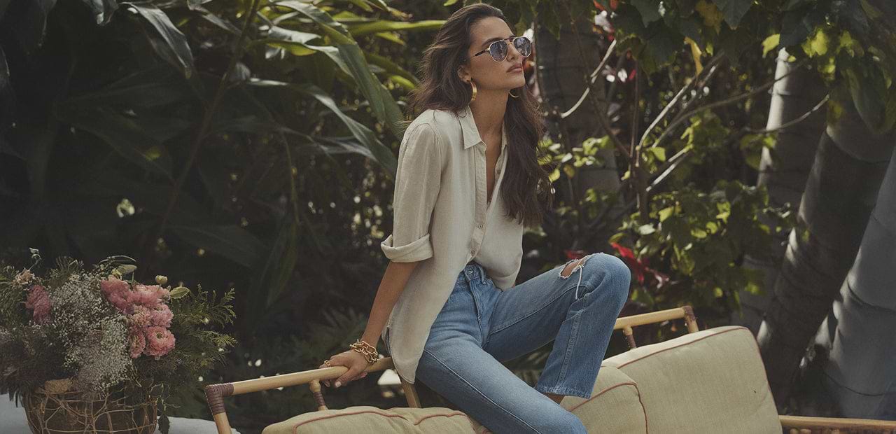 A woman wearing sunglasses, a white shirt, and blue jeans sits on an outdoor sofa surrounded by lush greenery and flowers, looking thoughtfully into the distance.