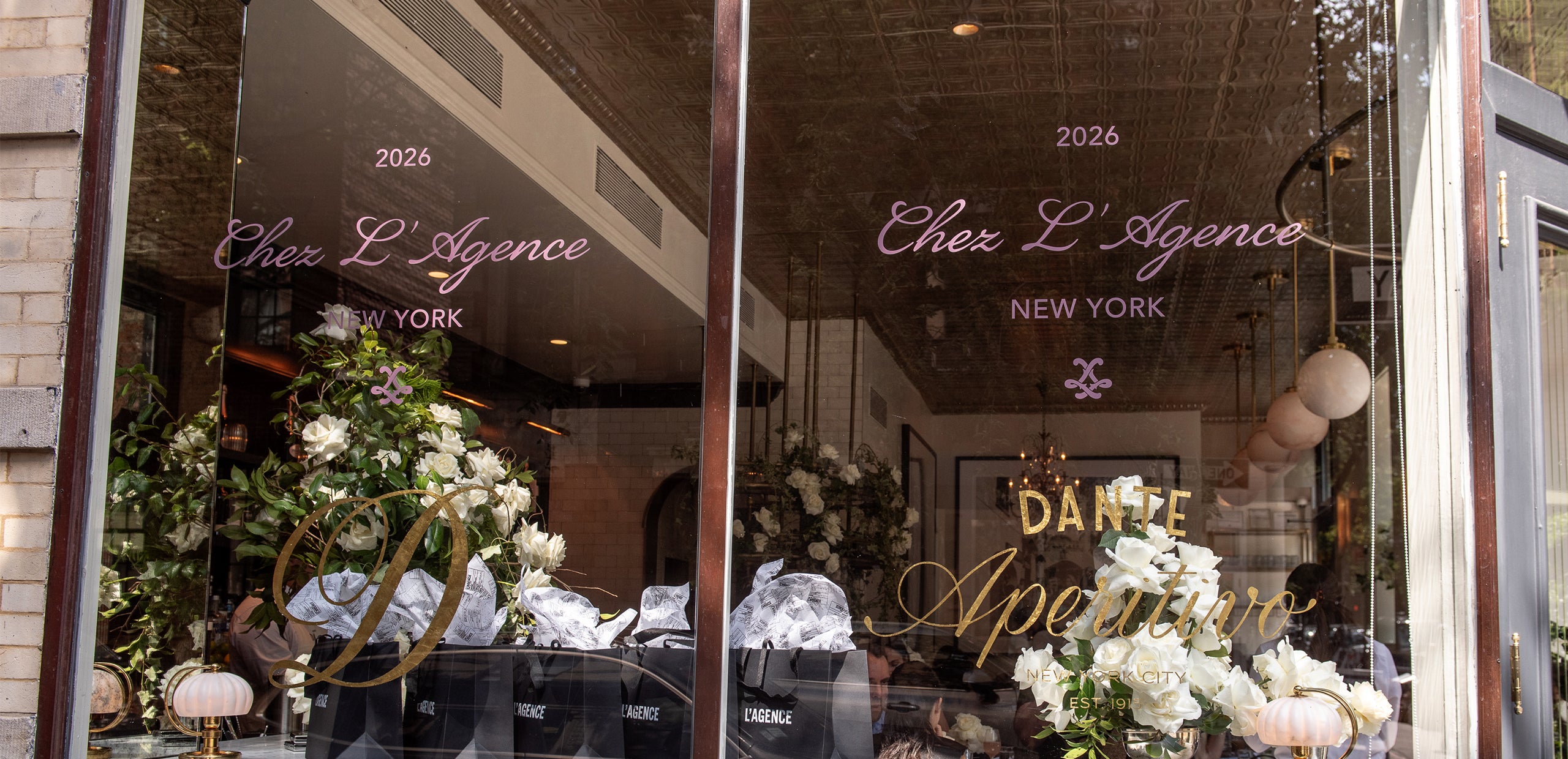Storefront window featuring Chez LAgence New York and Dante Aperitivo in gold and white script, with elegant flowers and bags displayed inside. The glass captures reflections of surrounding buildings and the city street.