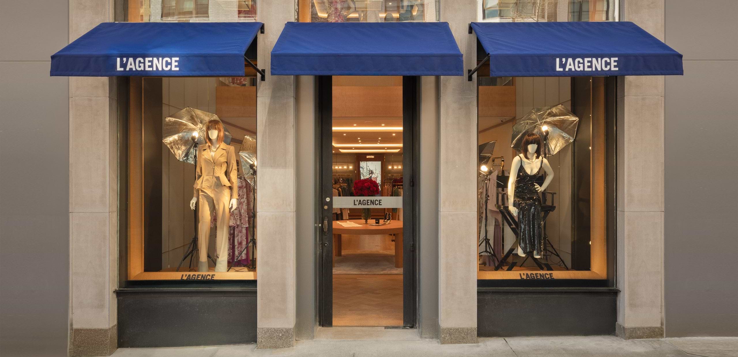 Storefront of LAGENCE with blue awnings, large display windows featuring mannequins in stylish outfits, and a glass door entrance in the center. The store interior is visible through the door and windows.