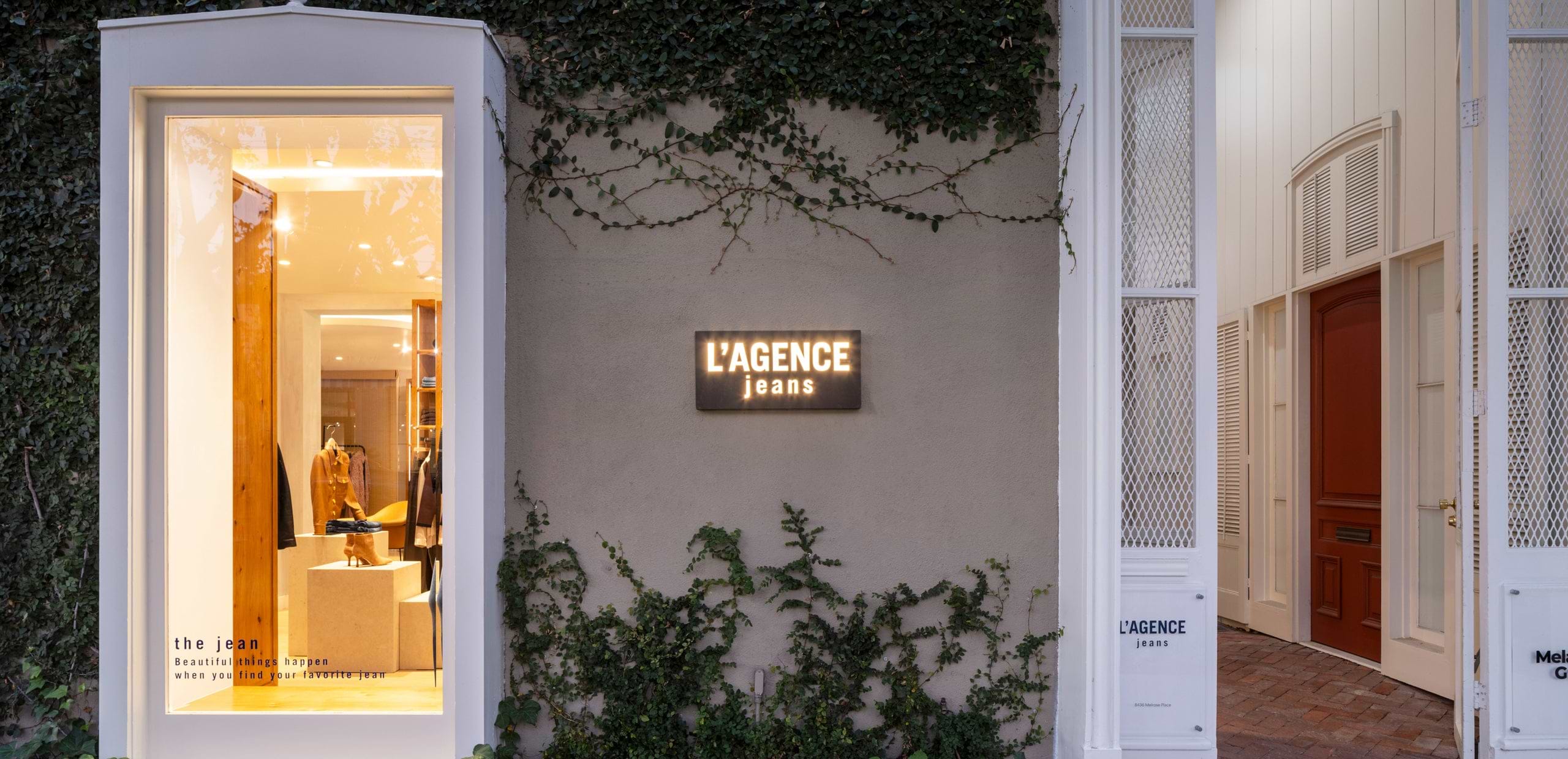 The exterior of the L’AGENCE jeans store features an illuminated sign on a beige wall with ivy, a large display window showing clothing inside, and an open white door leading into the shop.