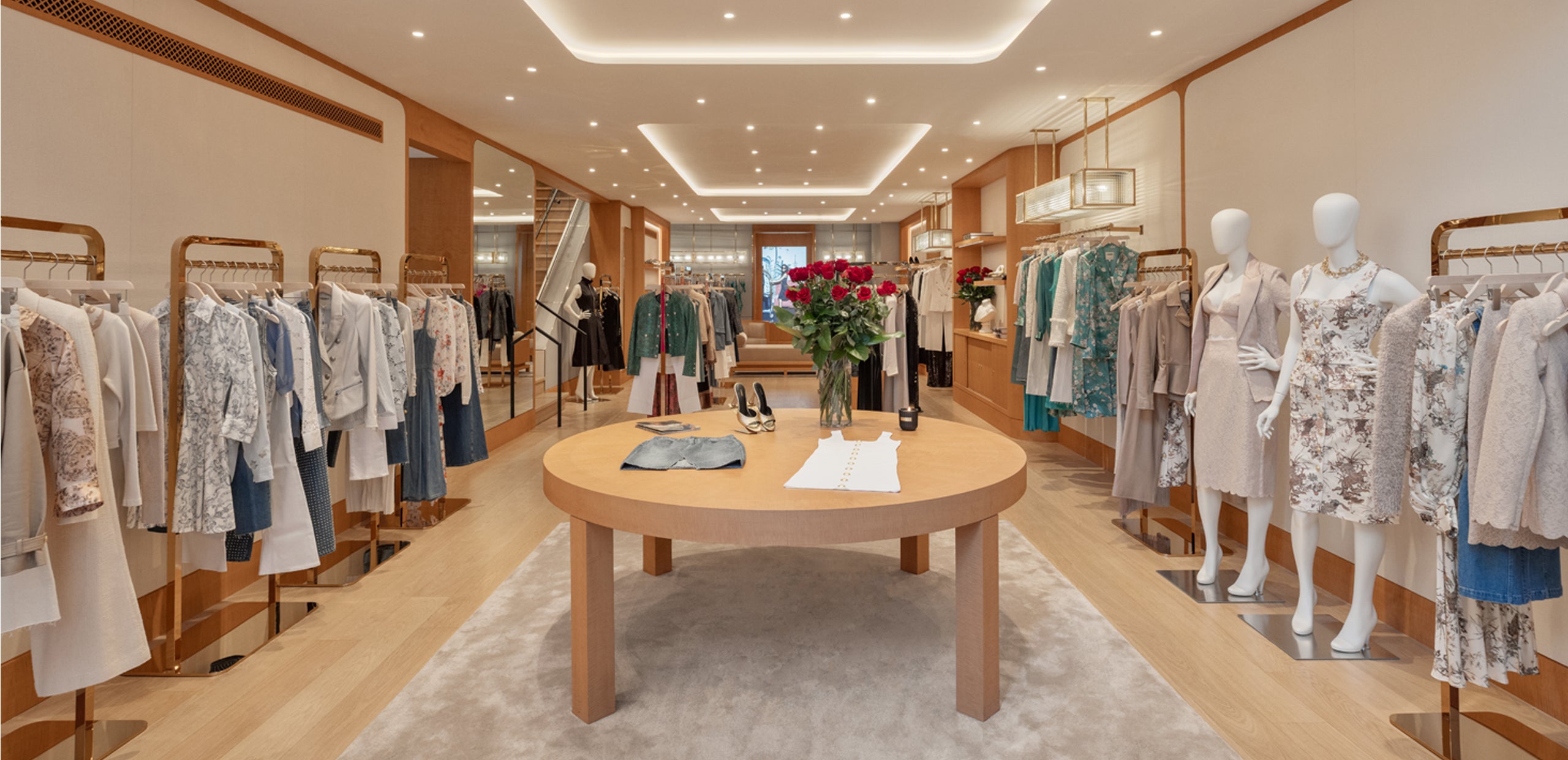 A modern clothing boutique with wooden floors, racks of dresses and blouses, mannequins in floral outfits, and a round table with flowers and accessories in the center under bright ceiling lights.
