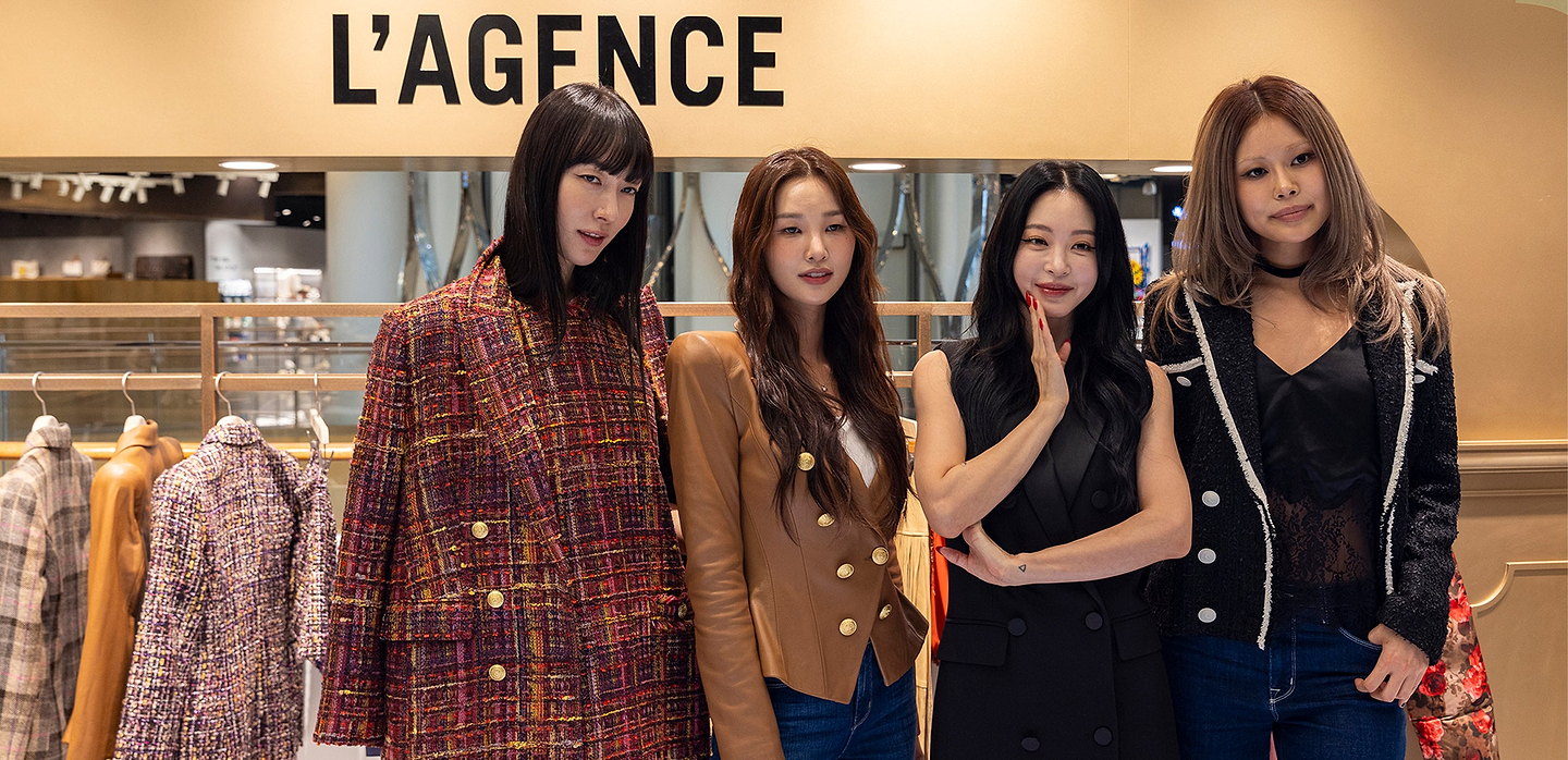 Four women pose together in stylish outfits in front of a clothing store display with the sign LAGENCE above them. The background features racks of colorful clothes and a bright, modern boutique interior.