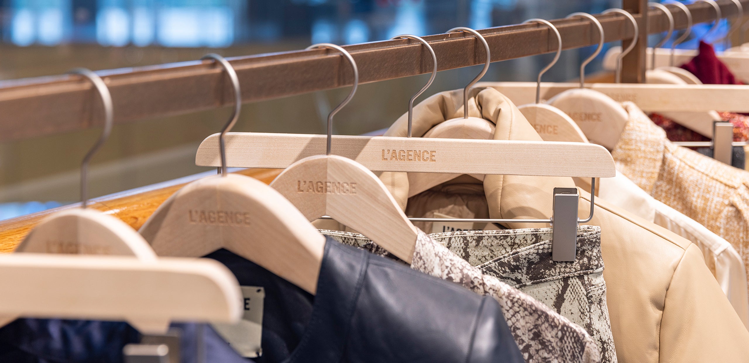 A row of wooden hangers labeled L’AGENCE holding various stylish jackets and coats, including patterned and solid options, hung neatly on a clothing rack in a store.