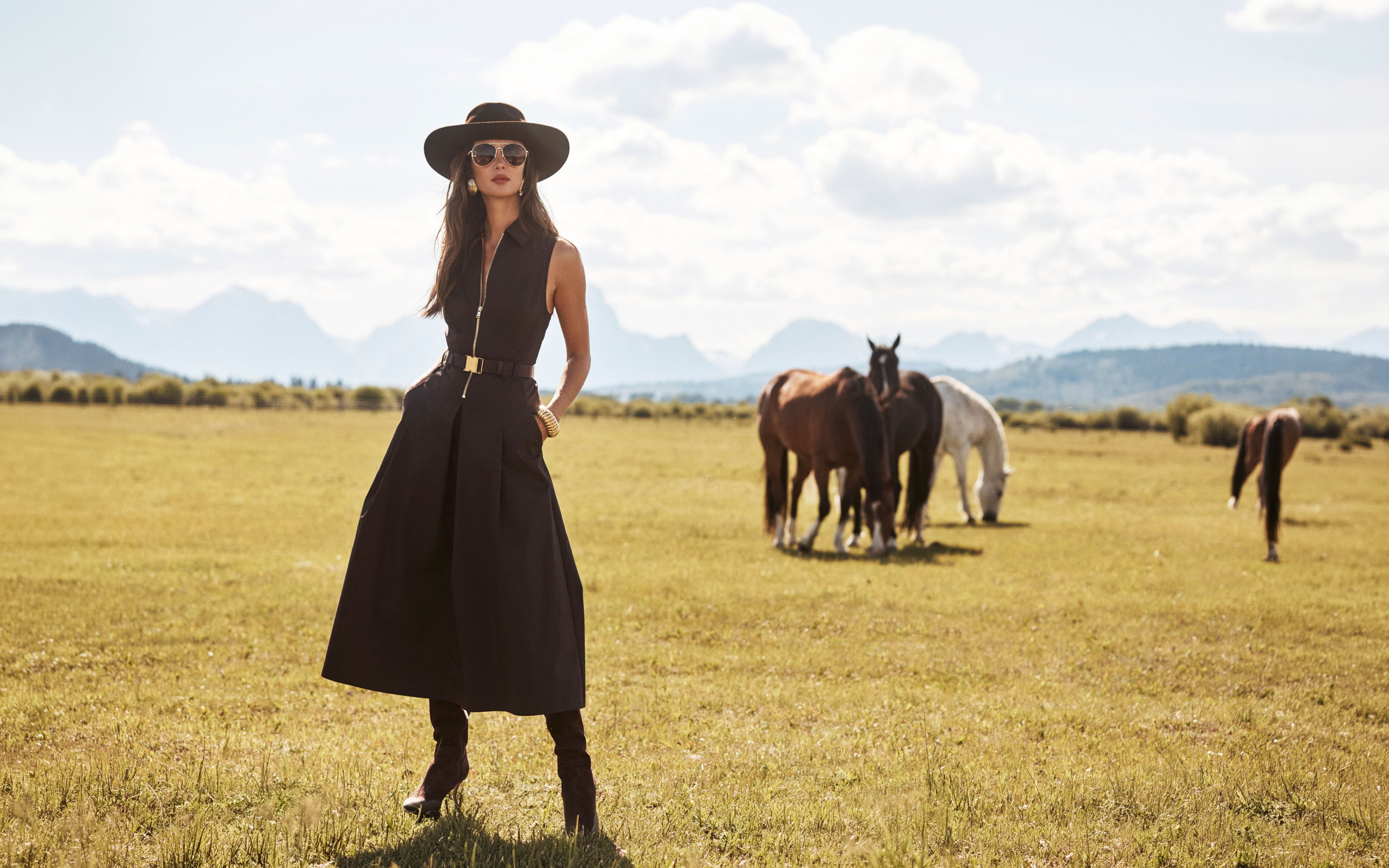 A woman in a black dress, boots, hat, and sunglasses stands in a grassy field with mountains in the background. Several horses graze behind her under a partly cloudy sky.