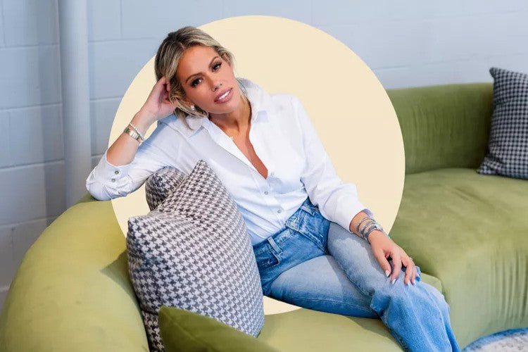 A woman with blonde hair, wearing a white shirt and blue jeans, sits casually on a green sofa with patterned pillows, smiling and resting her elbow on the backrest.