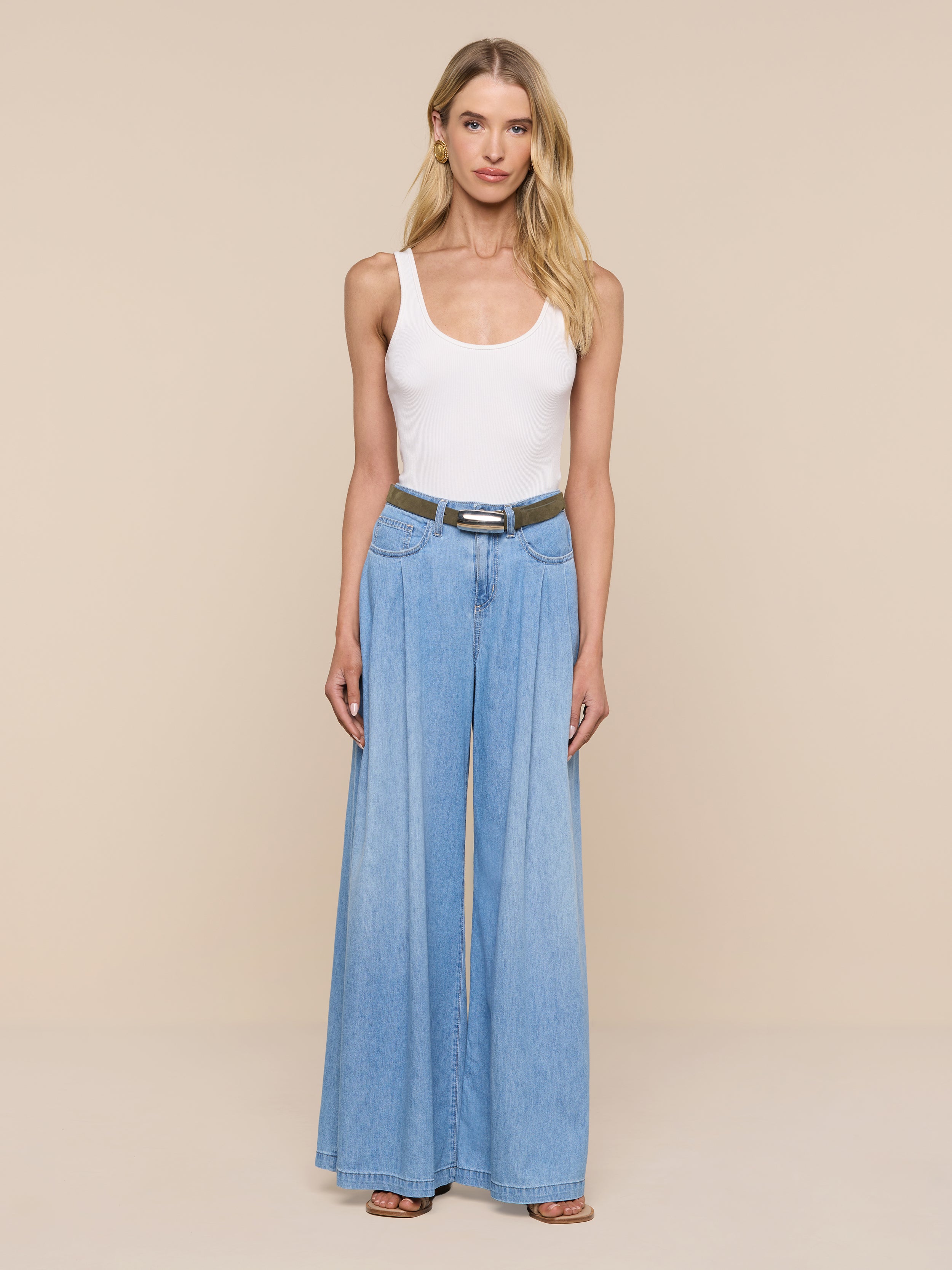 Una donna è in piedi su uno sfondo beige e indossa un top bianco senza maniche, jeans azzurri a gamba larga, sandali neri e la cintura in pelle scamosciata Marlowe color verde oliva con fibbia placcata argento, per un look minimalista.
