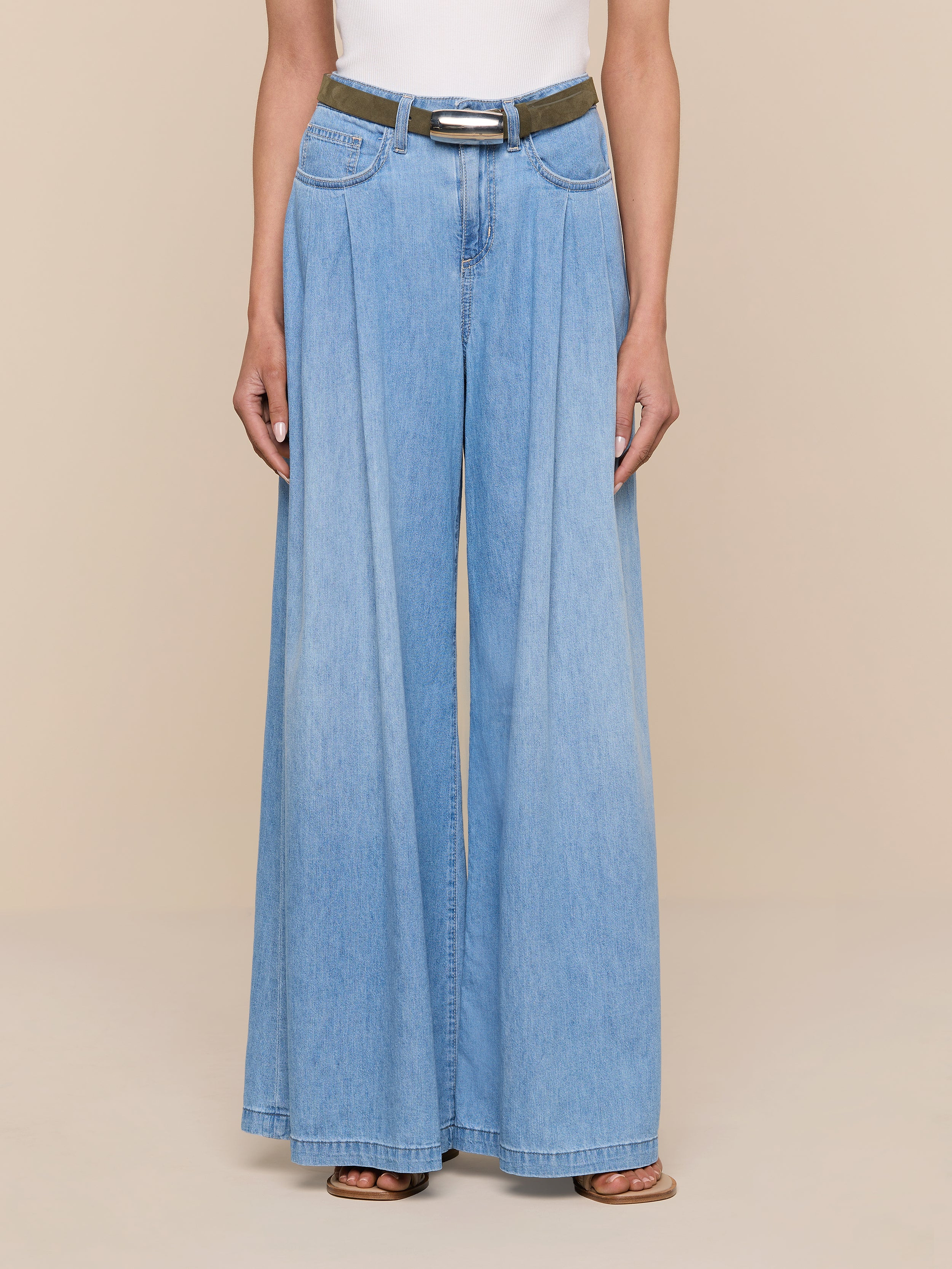 Una persona che indossa jeans in denim azzurro a gamba larga con pieghe, un top bianco, la cintura in pelle scamosciata Marlowe verde oliva con fibbia placcata argento e sandali marroni è in posa su uno sfondo beige: un perfetto esempio di stile minimalista.