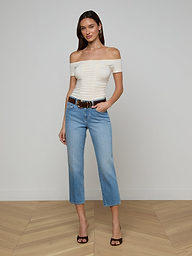 A woman stands against a plain background, smiling slightly with one hand in her pocket. She wears an off-the-shoulder white top, high-waisted blue cropped jeans, black heels, earrings, and the Tobar Suede Belt with a mixed-metal buckle.
