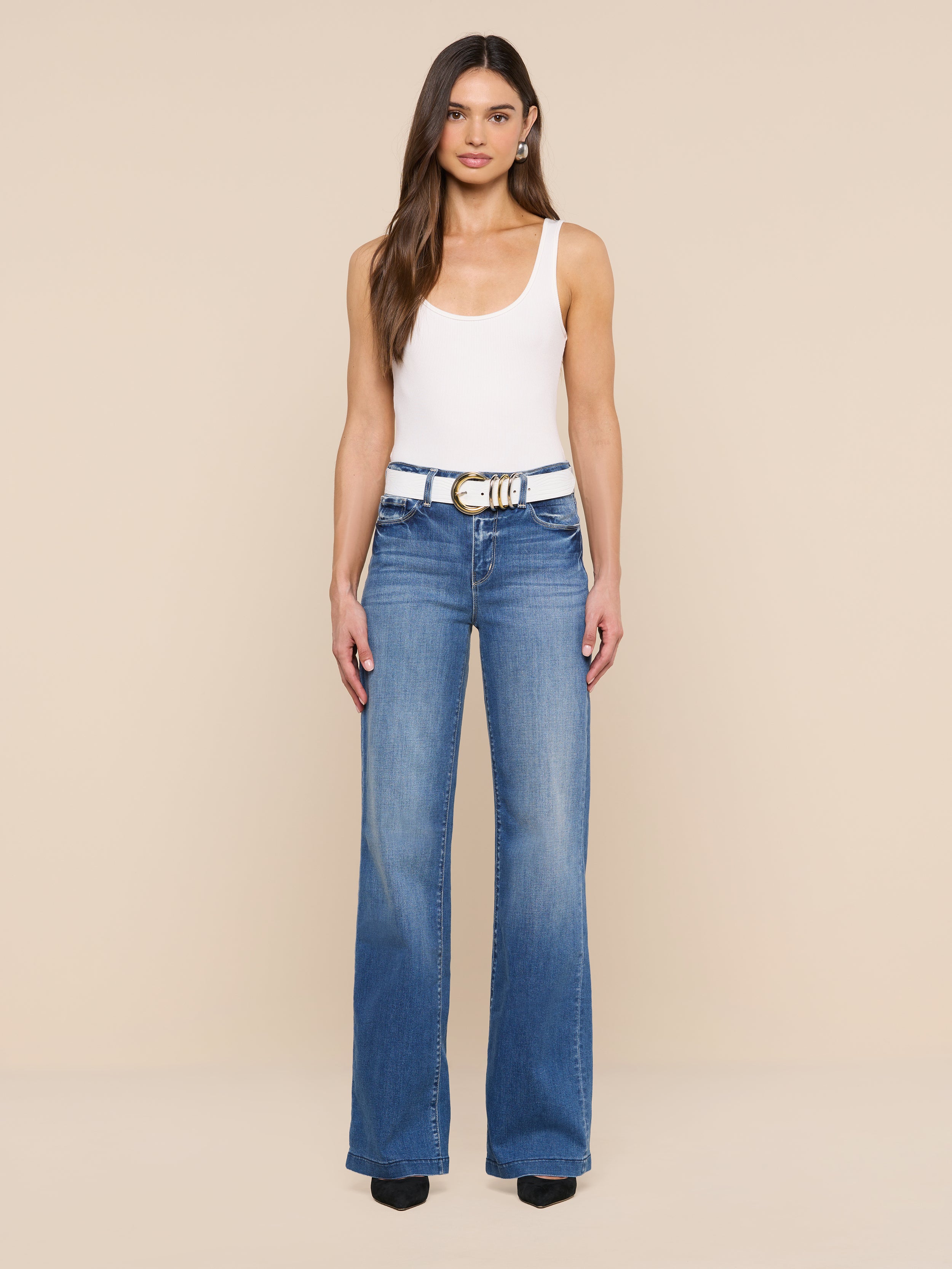 A woman stands facing forward, wearing a white tank top, blue high-waisted jeans, and black shoes. Her look is completed by the Tobar Leather Belt, featuring an embossed leather design and mixed-metal buckle, set against a plain white background.