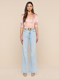 Una donna è ritratta di fronte su uno sfondo beige; indossa una camicetta di raso rosa chiaro, jeans a vita alta e a gamba larga di colore azzurro, la cintura Milou con fibbia in ottone e tacchi color carne.