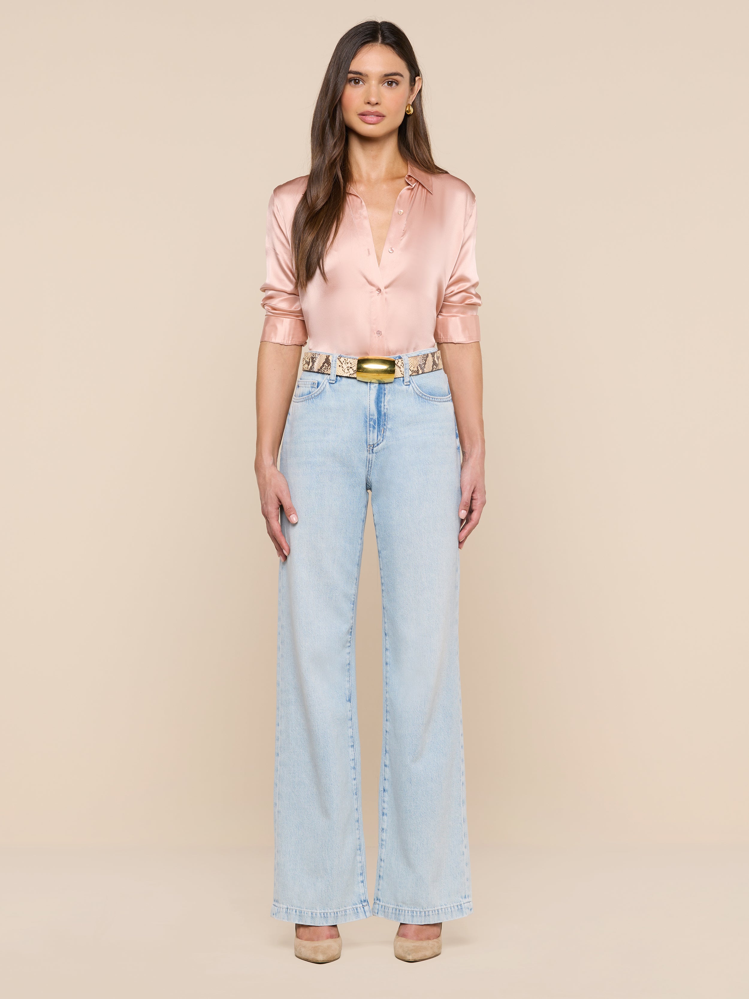 Una donna è ritratta di fronte su uno sfondo beige; indossa una camicetta di raso rosa chiaro, jeans a vita alta e a gamba larga di colore azzurro, la cintura Milou con fibbia in ottone e tacchi color carne.