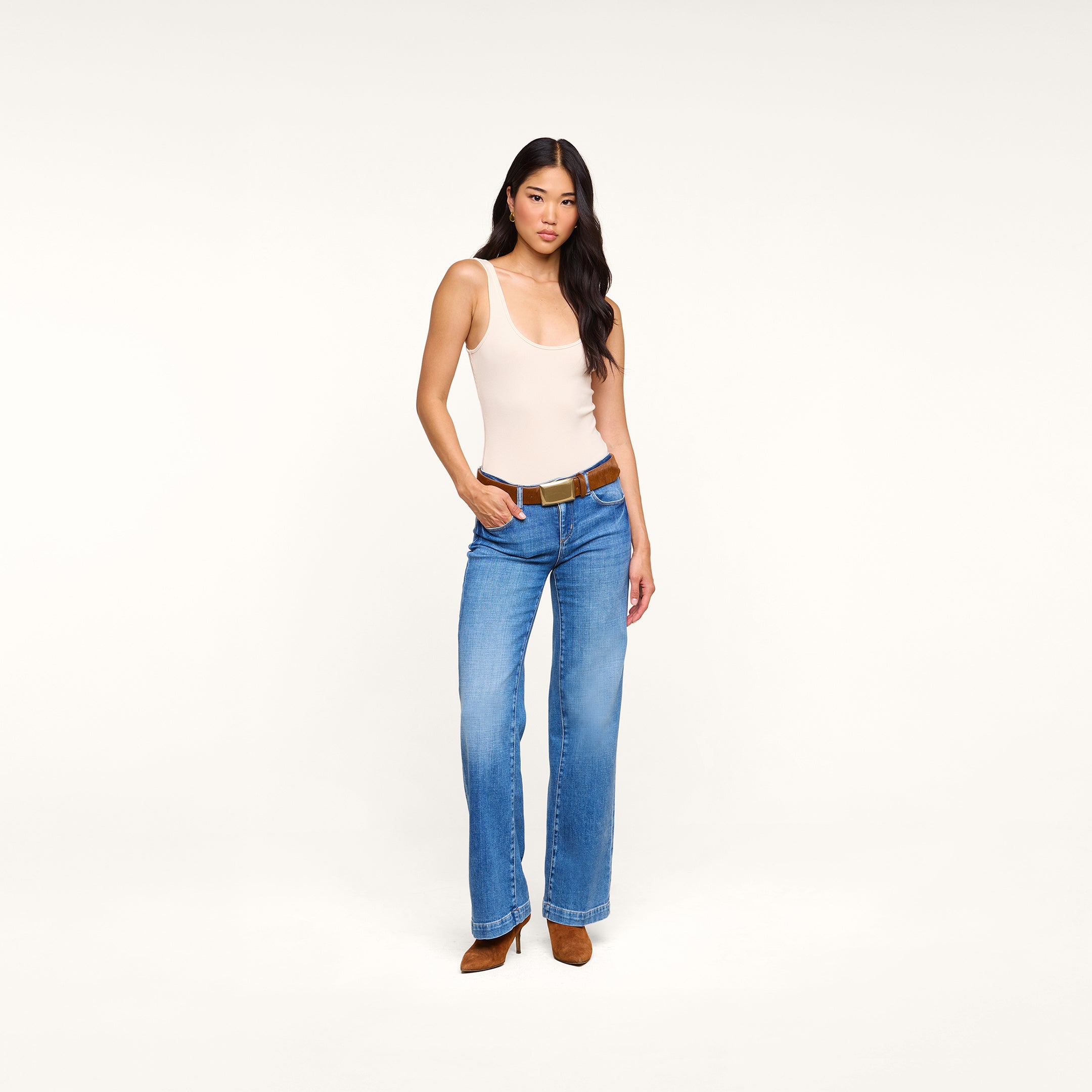 A woman with long dark hair and a neutral expression stands against a plain white background, wearing a sleeveless beige top, blue wide-leg jeans, brown shoes, and the handcrafted Sierra Belt—featuring a polished brass buckle—made in LA.