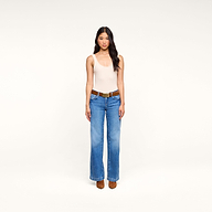 A woman stands against a plain white background, wearing a beige tank top, blue wide-leg jeans paired with the Sierra Belt featuring a polished brass buckle and calf hair leather, and brown shoes. She has long dark hair and a neutral expression.