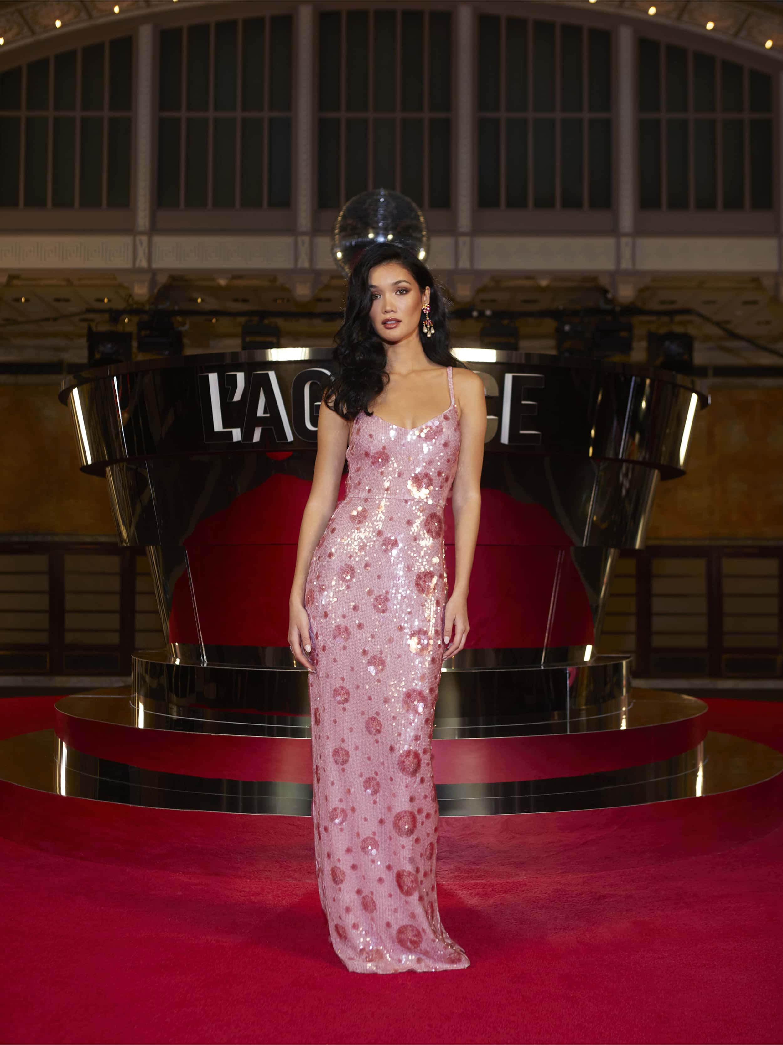 A woman in the Karma Sequin Maxi Dress, a shimmering pink gown, stands on a red carpet before a modern reflective backdrop with LAGENCE lit behind her. Her long dark hair and elegant posture are enhanced by the warm ambient lighting.