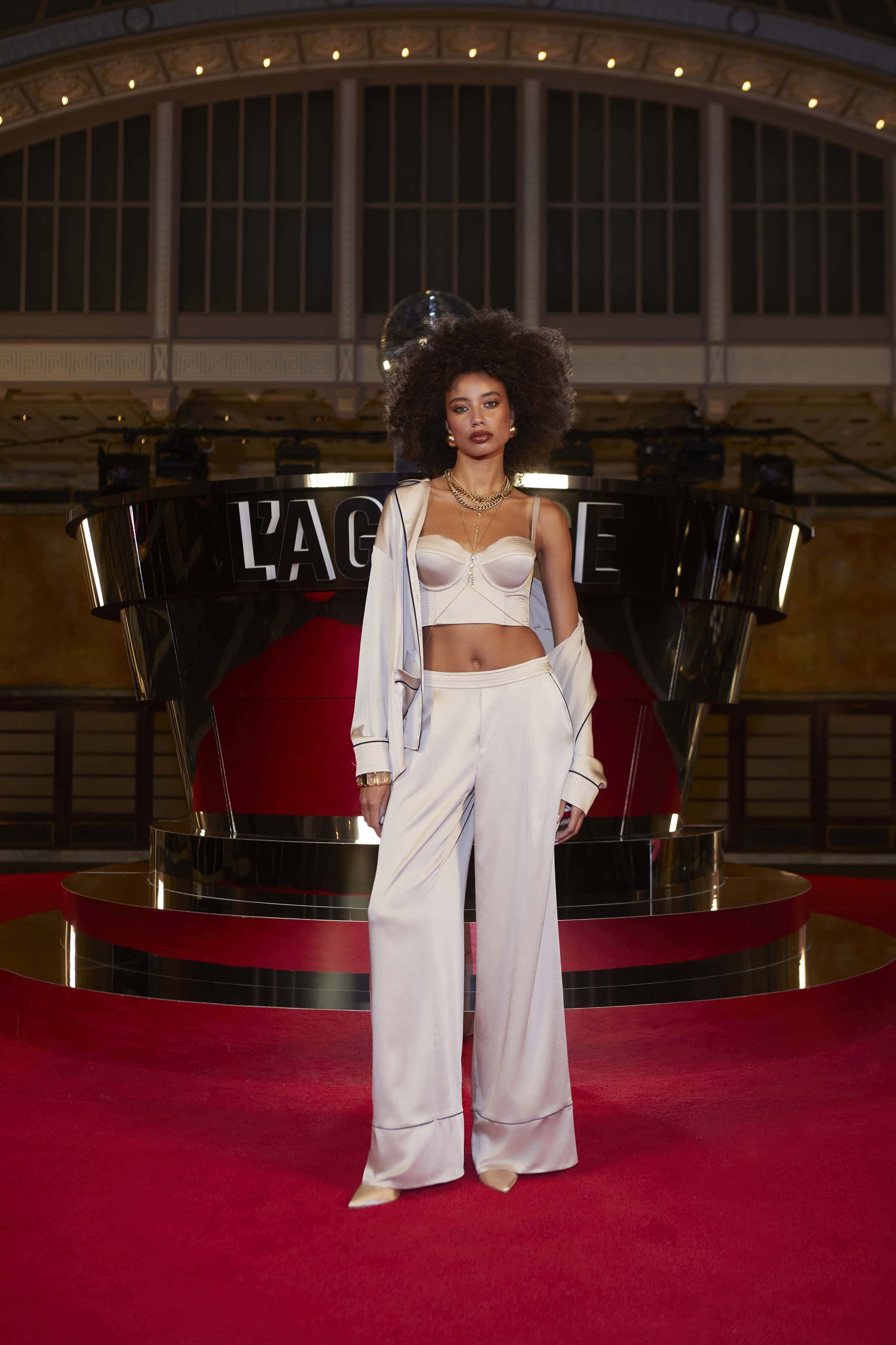 A woman with an afro hairstyle stands confidently on a red carpet, wearing the Tavia Trouser, a white bralette, and a loose jacket. Behind her is a shiny black and silver structure with large lettering.