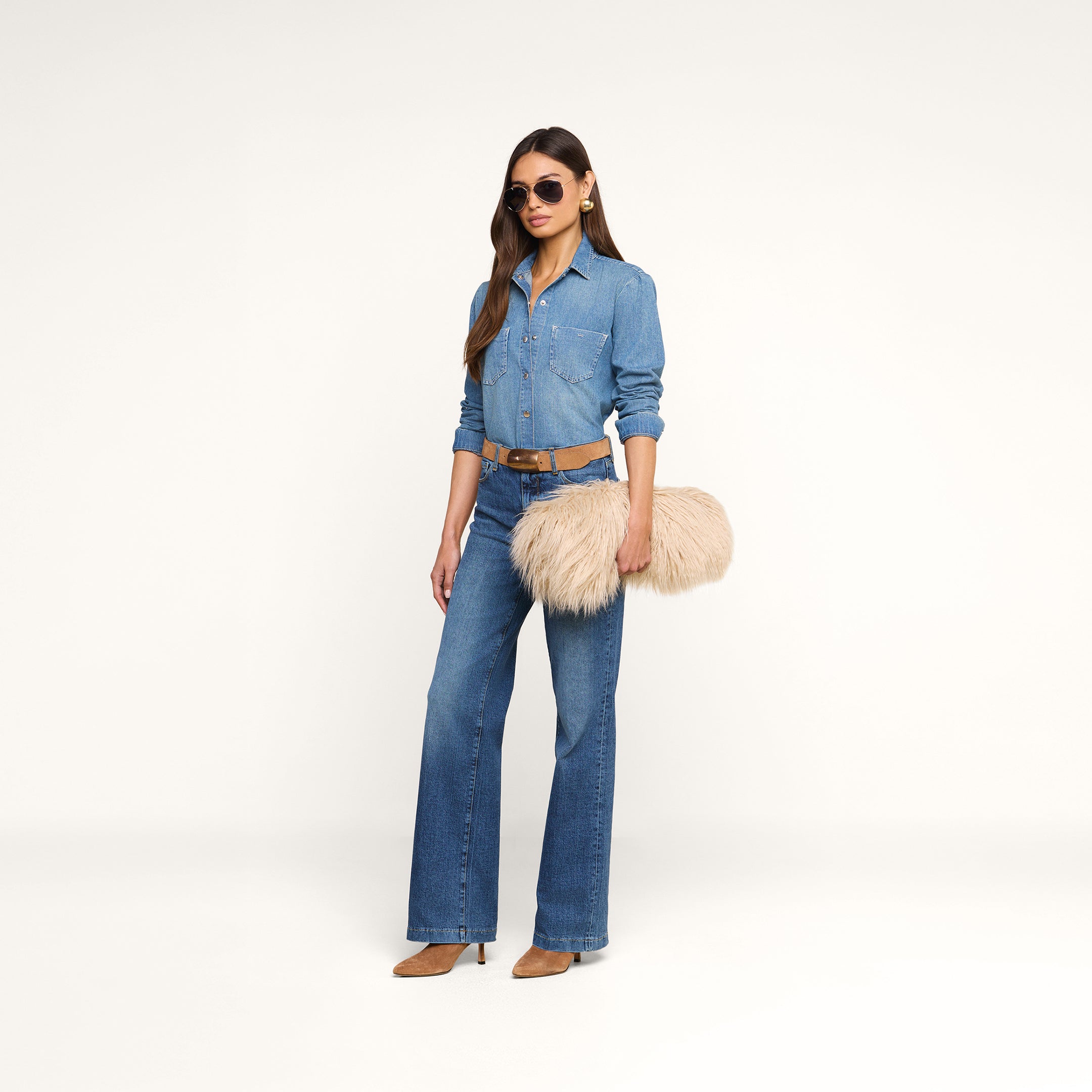 A woman in a denim shirt and flared jeans stands against a white background, wearing sunglasses and holding the Adelina Faux Fur Clutch. She completes her look with a brown belt and tan heeled shoes.