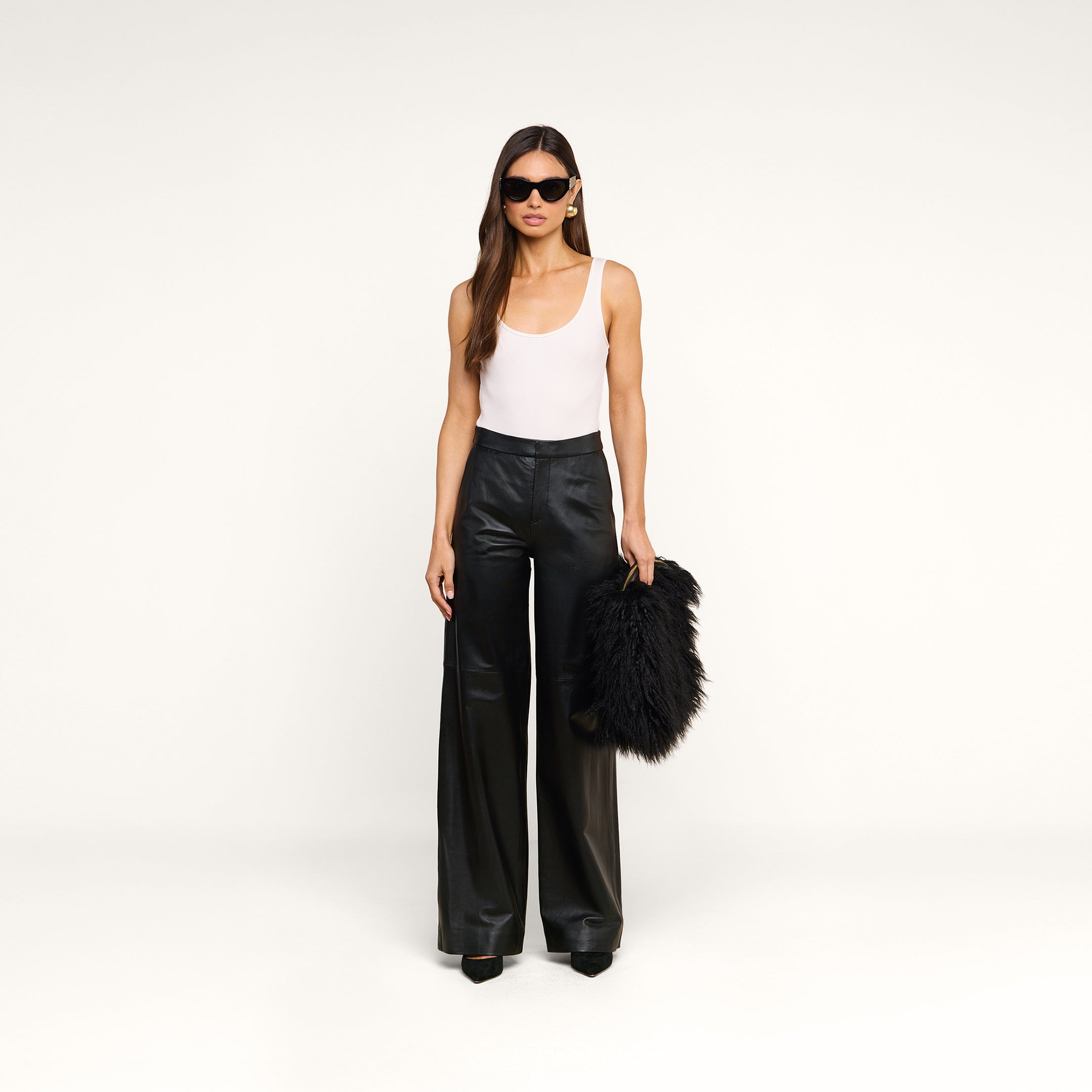 A woman stands against a plain background, wearing sunglasses, a white tank top, and black wide-leg pants while holding the Adelina Faux Fur Clutch in her left hand.