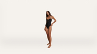 A woman stands barefoot on a white background, wearing a black one-piece swimsuit with thin straps and a cutout below the chest. She has long hair and one hand resting on her hip.