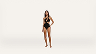 A woman stands barefoot wearing a black, asymmetrical one-piece swimsuit with cutout details, posing against a plain white background.