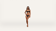 A woman stands barefoot on a plain white background, wearing a black bikini. She has long brown hair and her right hand rests on her hip while her left arm hangs by her side.
