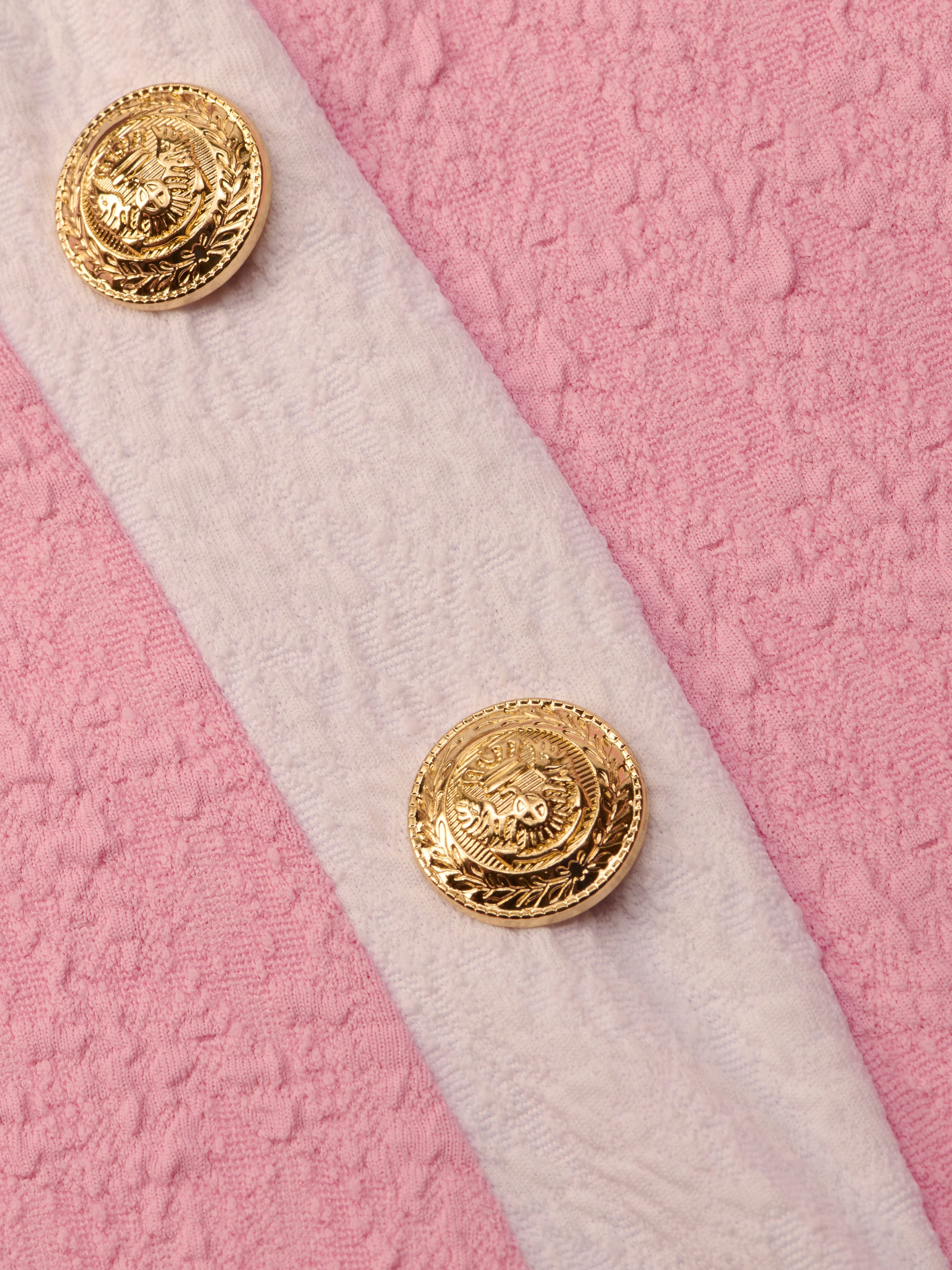 Close-up of two ornate gold buttons on the white bouclé fabric strap of the Tory Bandeau One-Piece Swimsuit, set against a textured pink background.
