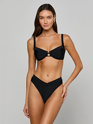A woman stands against a plain, light background wearing the Adrienne Bikini Top—a black bandeau-style top with underwire and Trapunto stitching—paired with a V-shaped bikini bottom.