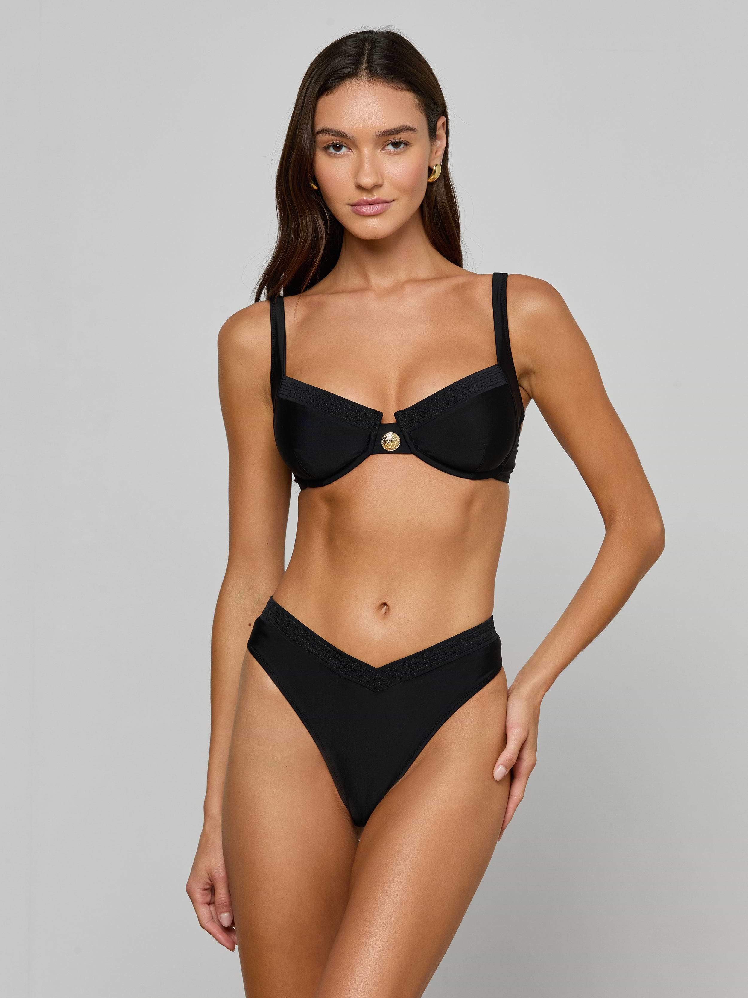 A woman stands against a plain, light background wearing the Adrienne Bikini Top—a black bandeau-style top with underwire and Trapunto stitching—paired with a V-shaped bikini bottom.