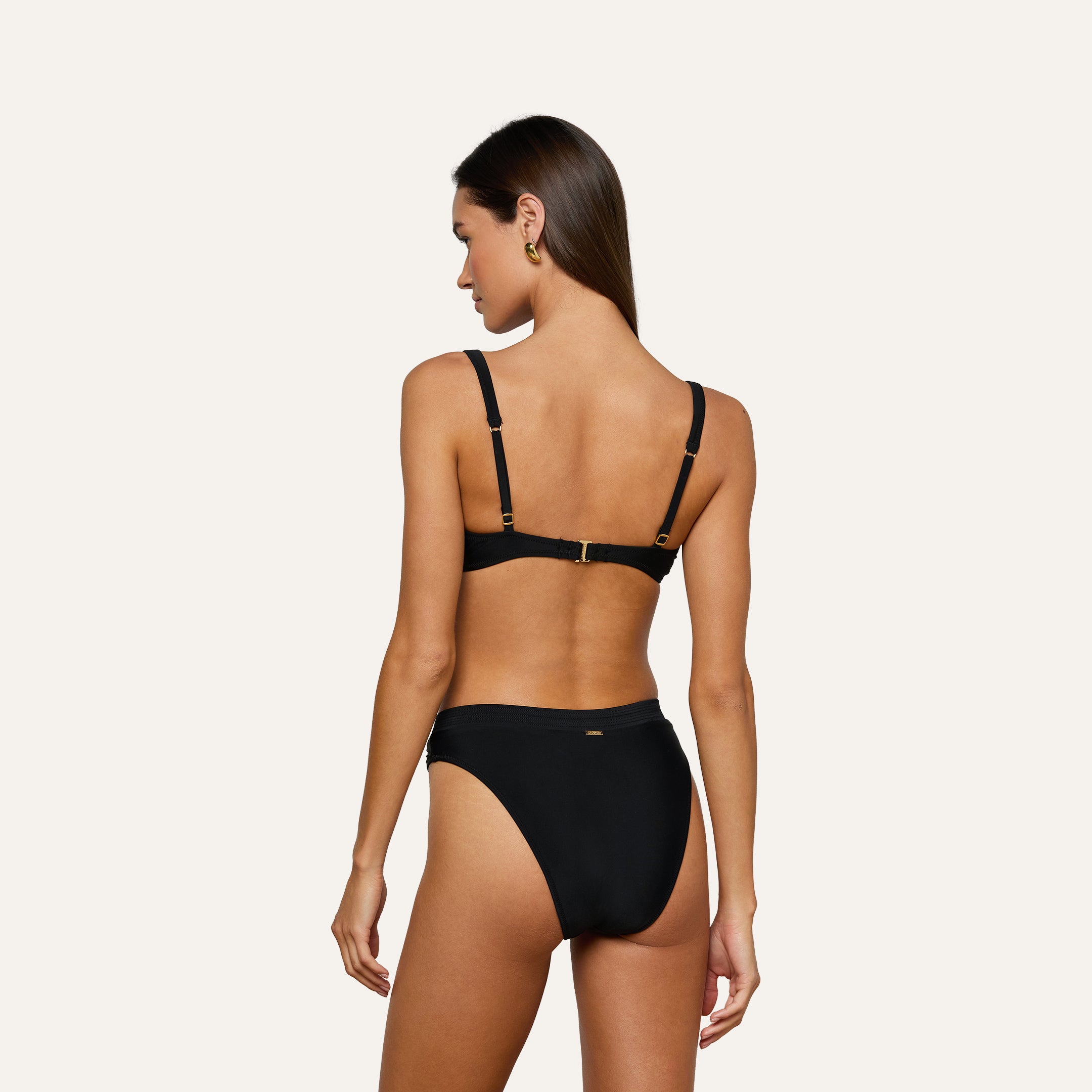 A woman with long brown hair is shown from the back, wearing the Adrienne Bikini Top—a black balconette style featuring gold accents and Trapunto stitching. She stands against a plain, light-colored background.