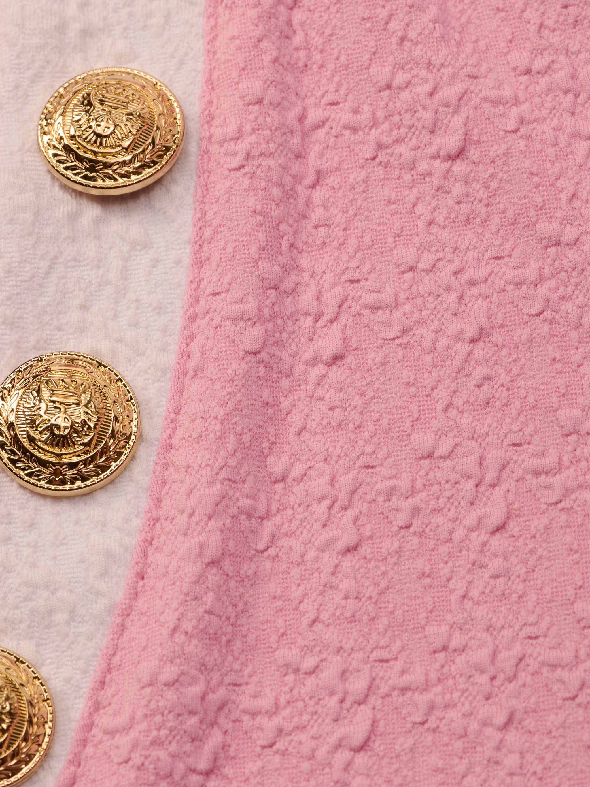 Four ornate gold buttons with engraved details are sewn onto light pink, water-safe bouclé fabric, arranged vertically along the edge of the Anastasia Cropped Rashguard.