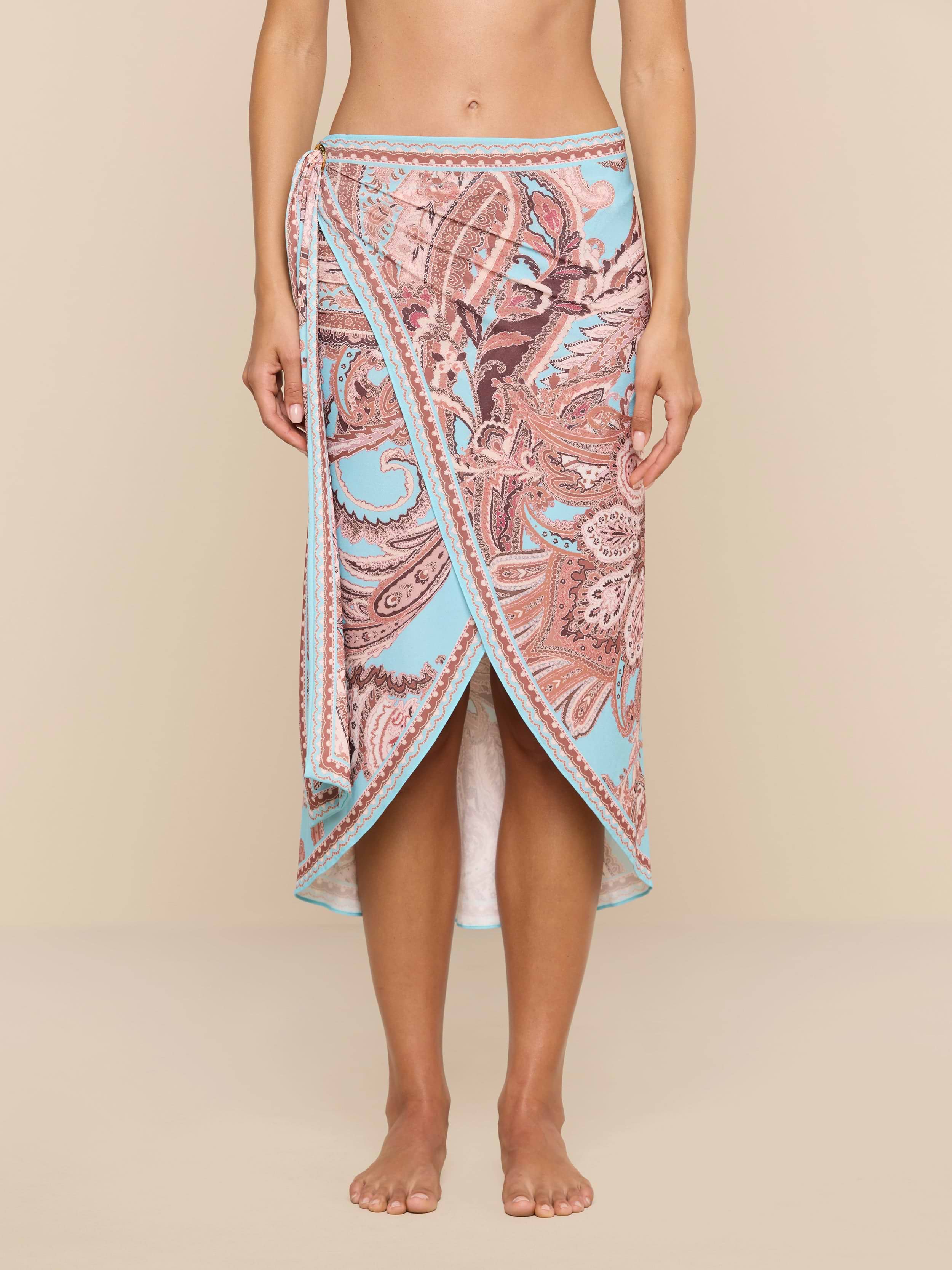 Barefoot against a plain white background, a person showcases the Marena Sarong in light blue and pink paisley print, embodying effortless beachside sophistication.