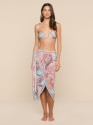 A woman stands barefoot against a plain white background, exuding beachside sophistication in a patterned bikini top and the Marena Sarong, which features matching blue and pink designs.
