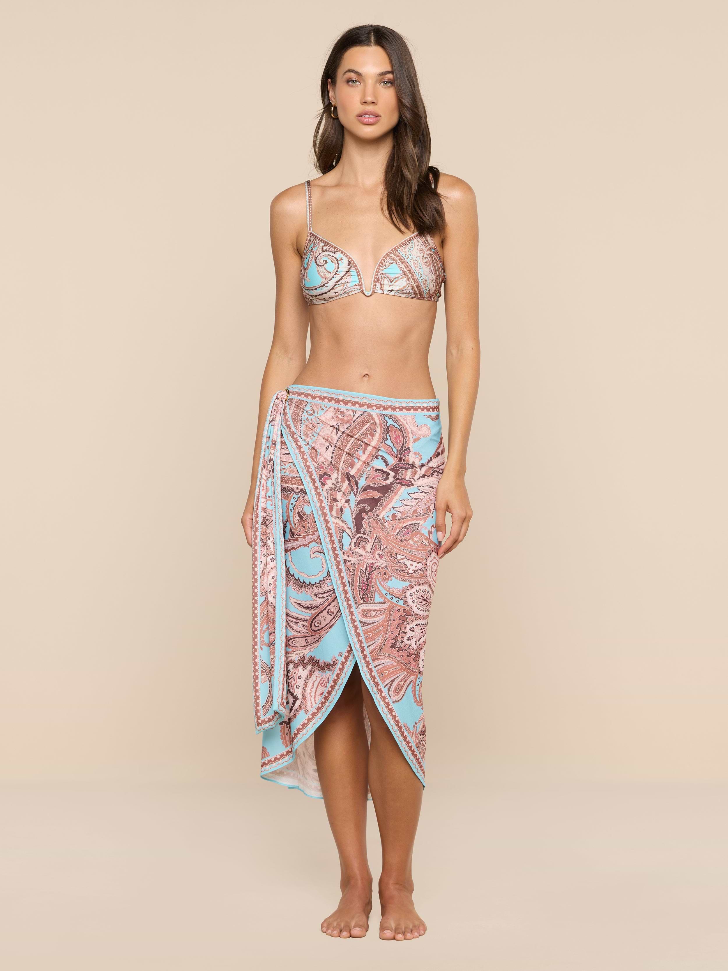 A woman stands barefoot against a plain white background, exuding beachside sophistication in a patterned bikini top and the Marena Sarong, which features matching blue and pink designs.