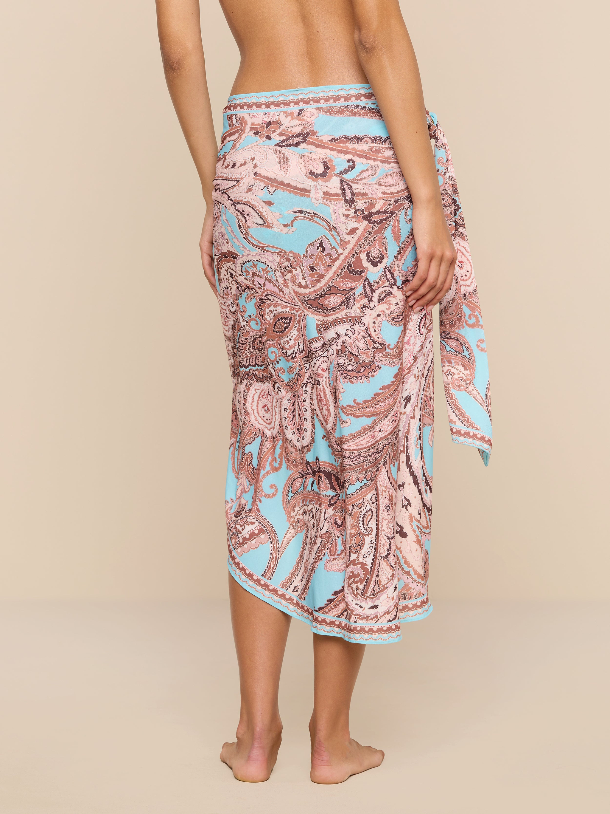 A person stands barefoot with their back to the camera, showcasing the Marena Sarong in a pink and blue paisley pattern tied at the waist. The upper body is partially visible, while the plain white background emphasizes this swimwear cover-up’s beachside sophistication.