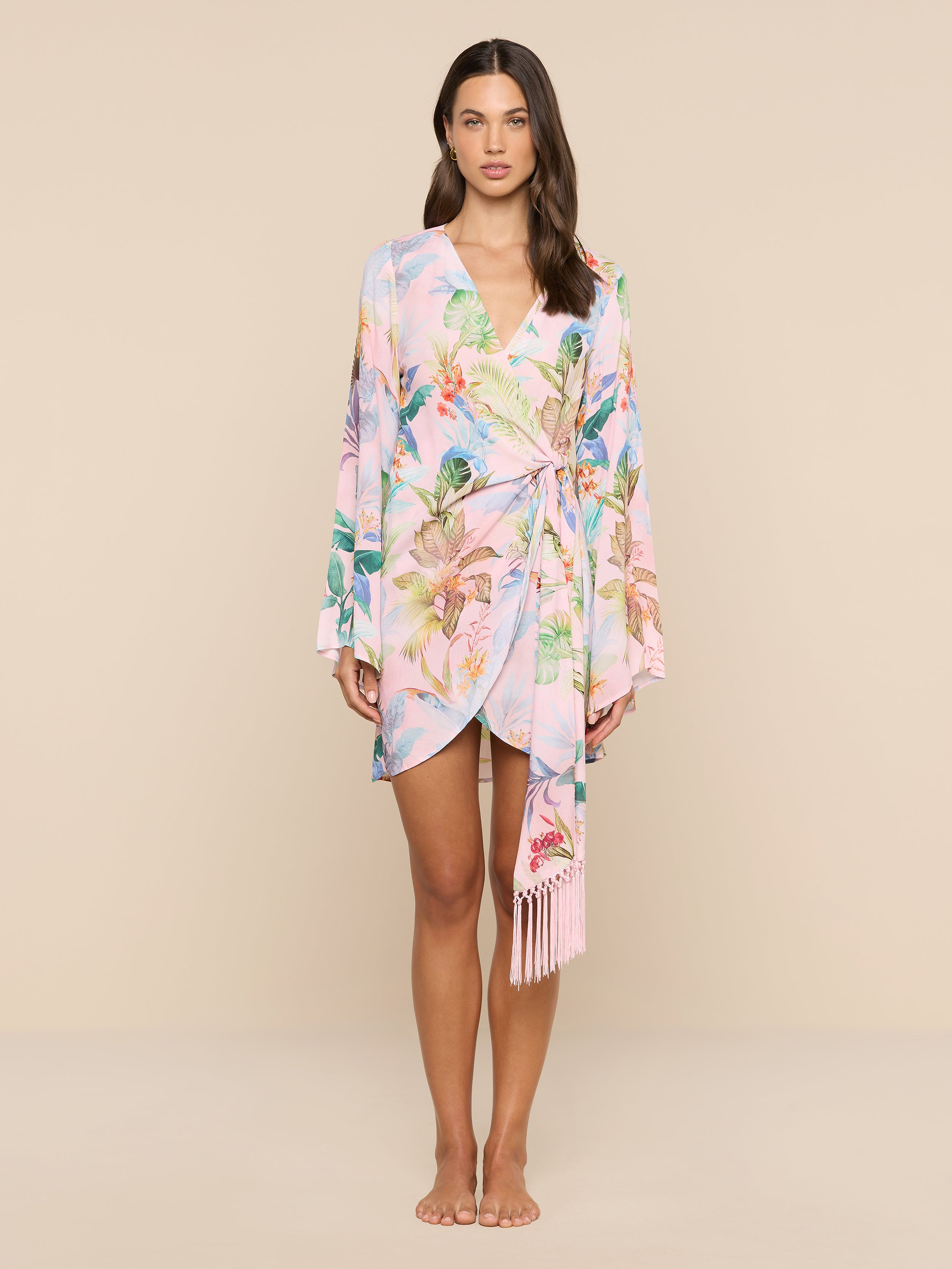 A woman stands facing forward, wearing the Kohima Cover-Up Dress—a pastel floral wrap dress with bell sleeves and a tie at the waist—against a plain white background.