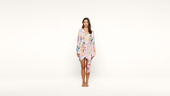 A woman stands barefoot against a plain white background, wearing a light pink floral kimono-style wrap dress with long sleeves and a tied waist.