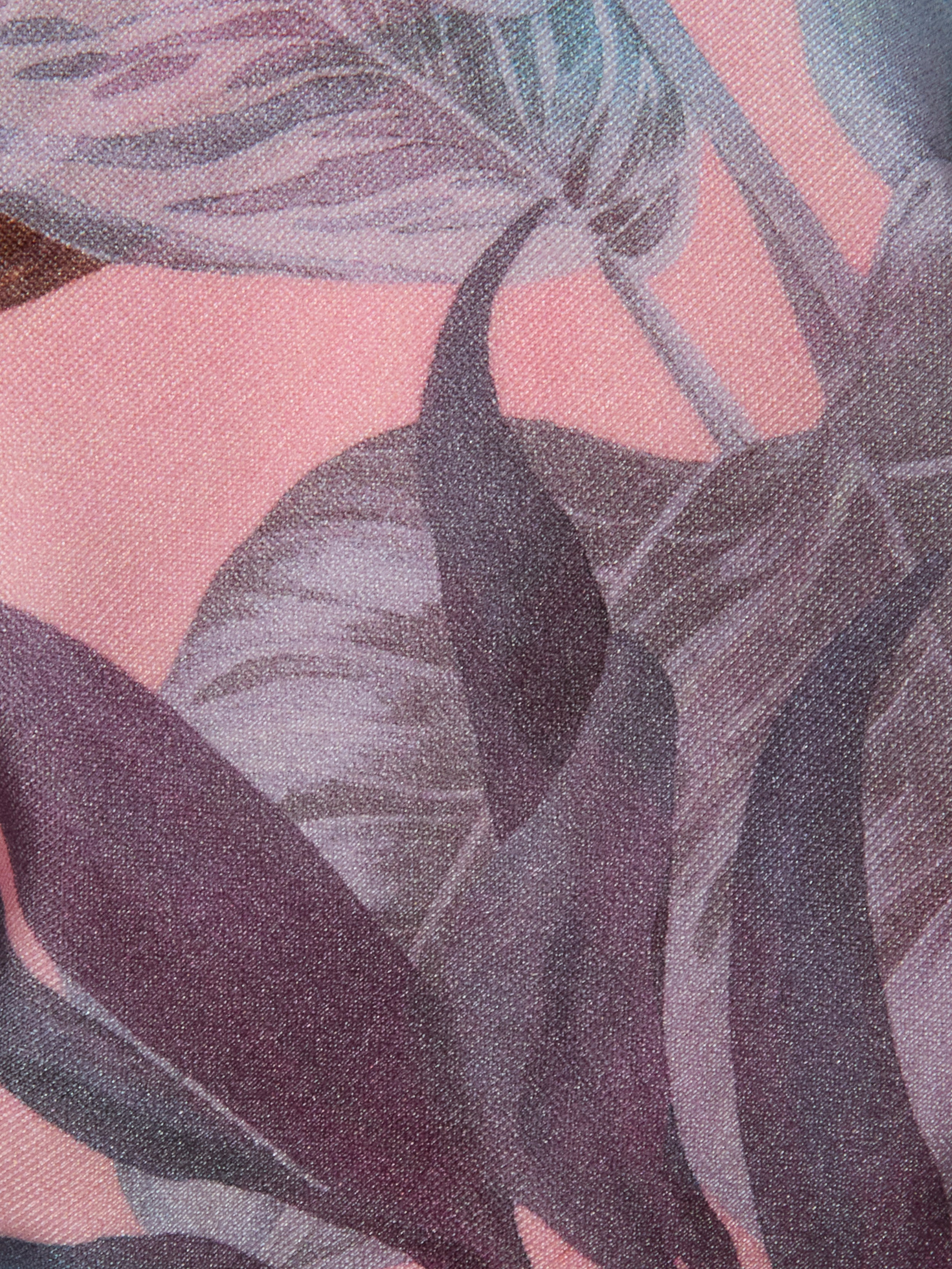 Close-up of the Suki Halter Bikini Top fabric, featuring a pink background with a printed pattern of large, overlapping purple and gray palm-like leaves. The texture of the fabric weave is visible.