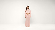 A woman stands barefoot against a white background, wearing a light pink, long-sleeved crop top and a matching long skirt. She has long, brown hair with bangs and a neutral expression.