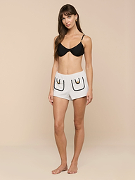 A woman stands barefoot against a plain background, wearing a black bra and the Amiya Cover-Up Short in light gray terrycloth, featuring two outlined pockets and black detailing.