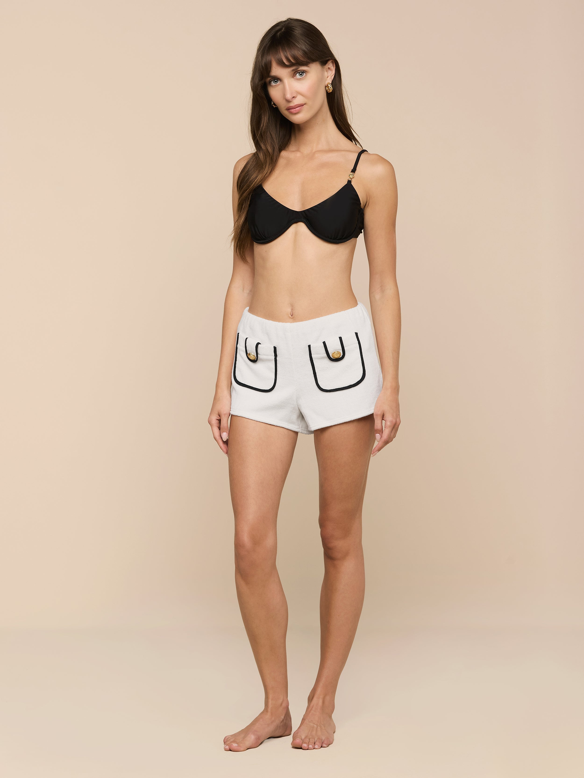 A woman stands barefoot against a plain background, wearing a black bra and the Amiya Cover-Up Short in light gray terrycloth, featuring two outlined pockets and black detailing.