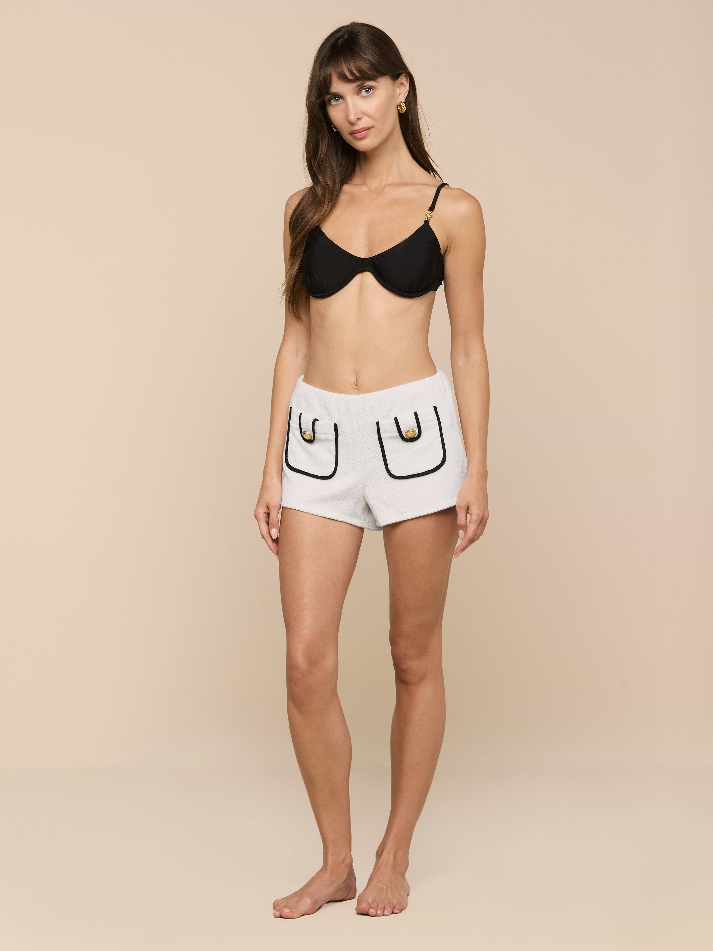 A woman stands barefoot against a plain background, wearing a black bra and the Amiya Cover-Up Short in light gray terrycloth, featuring two outlined pockets and black detailing.