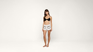 A woman stands barefoot against a plain white background, wearing a black bra and white shorts with black outlined pocket designs.