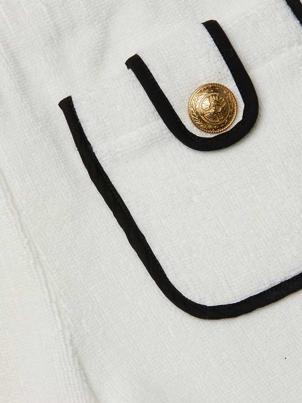 Close-up of a white textured Amiya Cover-Up Short pocket with black trim and a decorative gold button featuring an embossed design, reminiscent of classic terrycloth shorts perfect for poolside lounging.