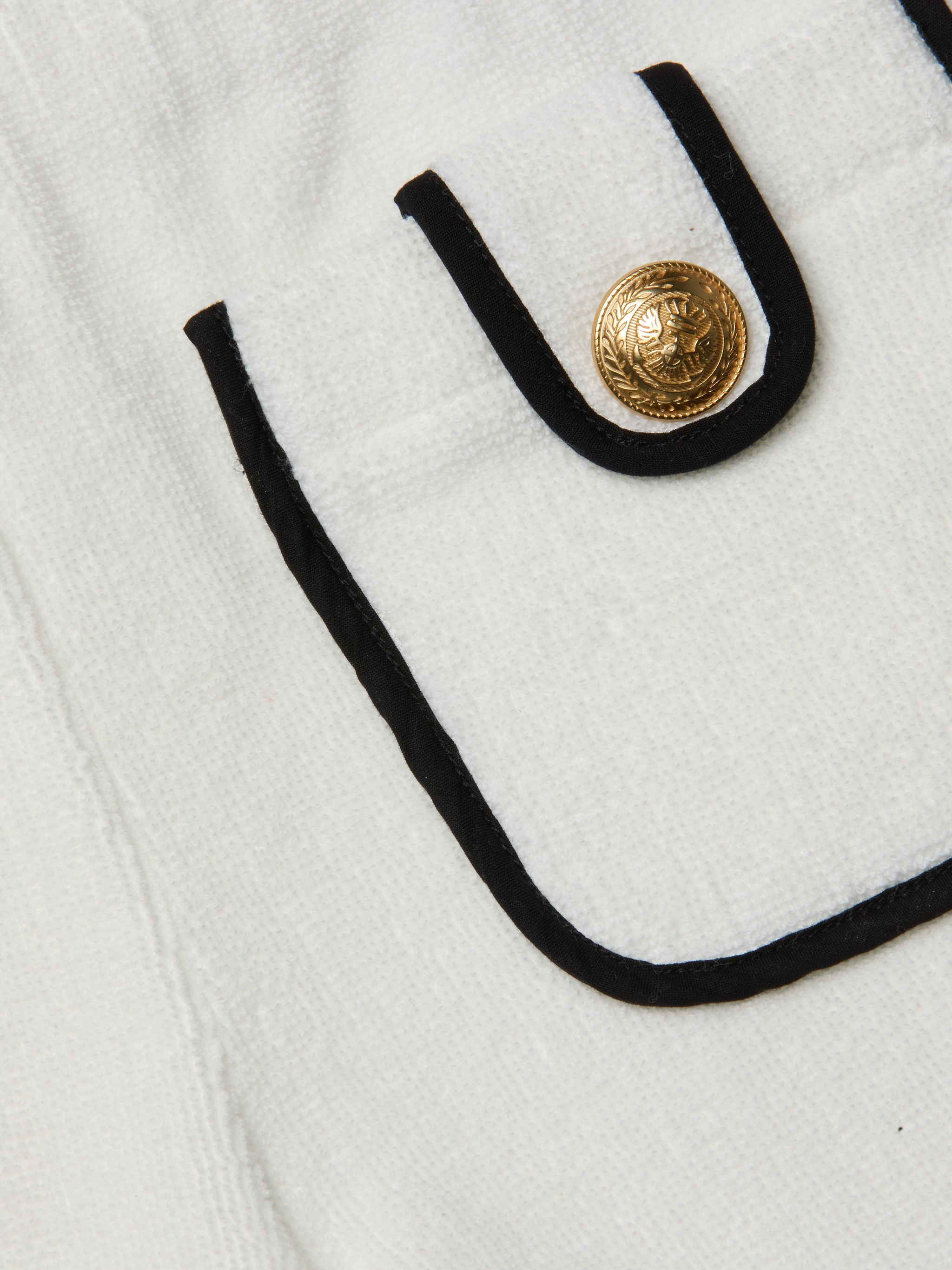 Close-up of a white textured Amiya Cover-Up Short pocket with black trim and a decorative gold button featuring an embossed design, reminiscent of classic terrycloth shorts perfect for poolside lounging.