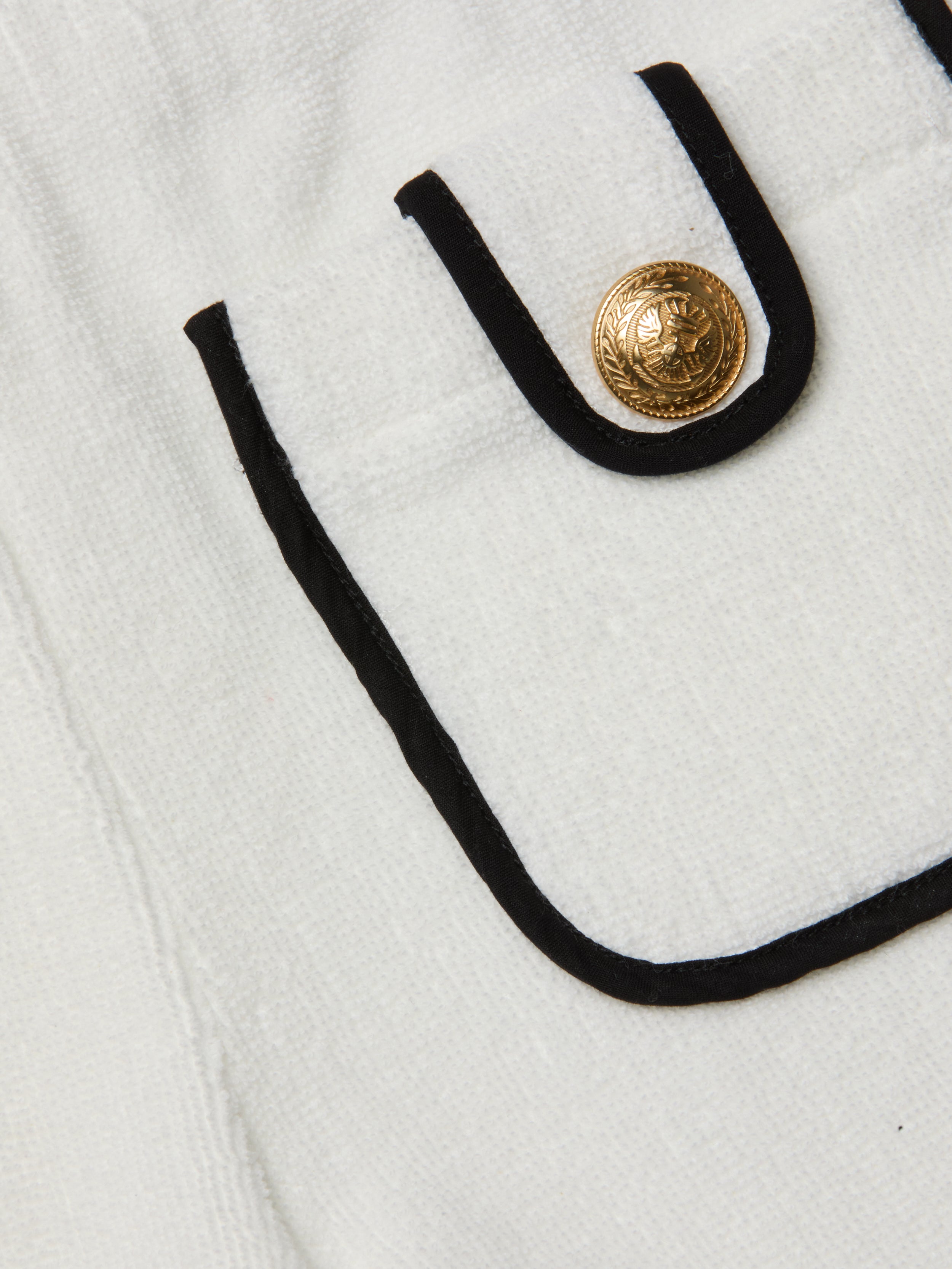 Close-up of a white textured Amiya Cover-Up Short pocket with black trim and a decorative gold button featuring an embossed design, reminiscent of classic terrycloth shorts perfect for poolside lounging.